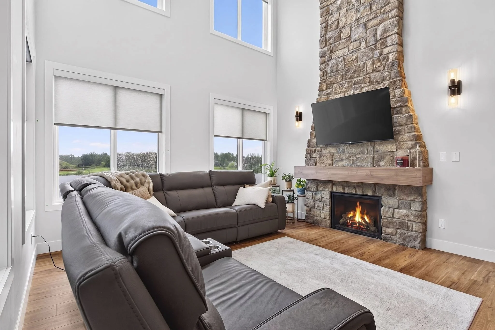 Living room with gray leather sofas, a stone fireplace with a mounted TV, and large windows showing a green outdoor landscape.