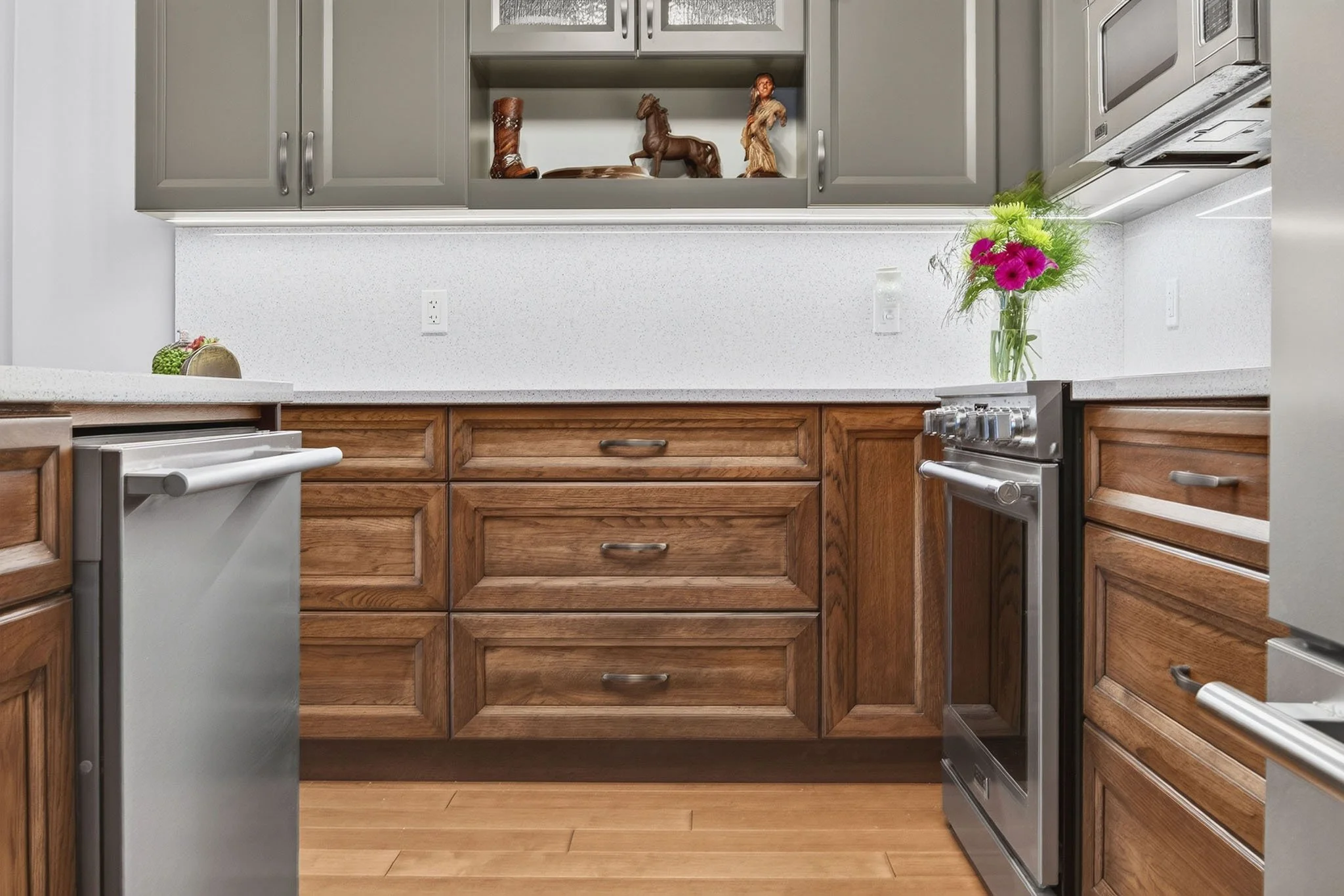 A kitchen with gray upper cabinets, wooden lower cabinets, a light-colored speckled countertop, and a hardwood floor. A stained glass vase of pink flowers is on the counter, and decorative statuettes of a cowboy boot, horse, and Native American woman
