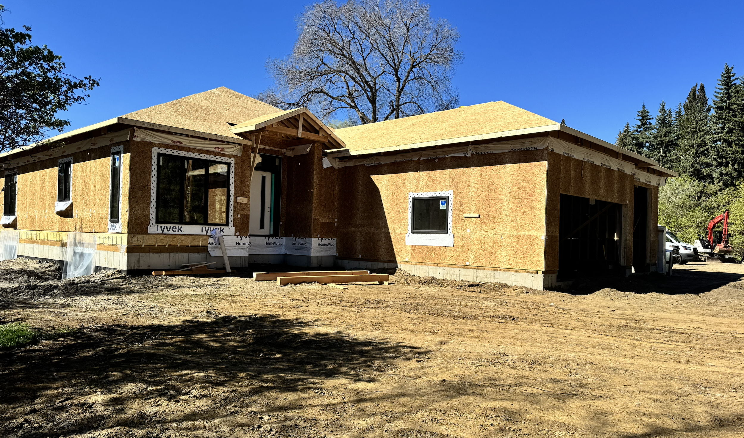 An acreage estate home under construction with plywood sheathing, three windows, and a gabled roof, set against a bright blue sky with a large leafless tree in the background.