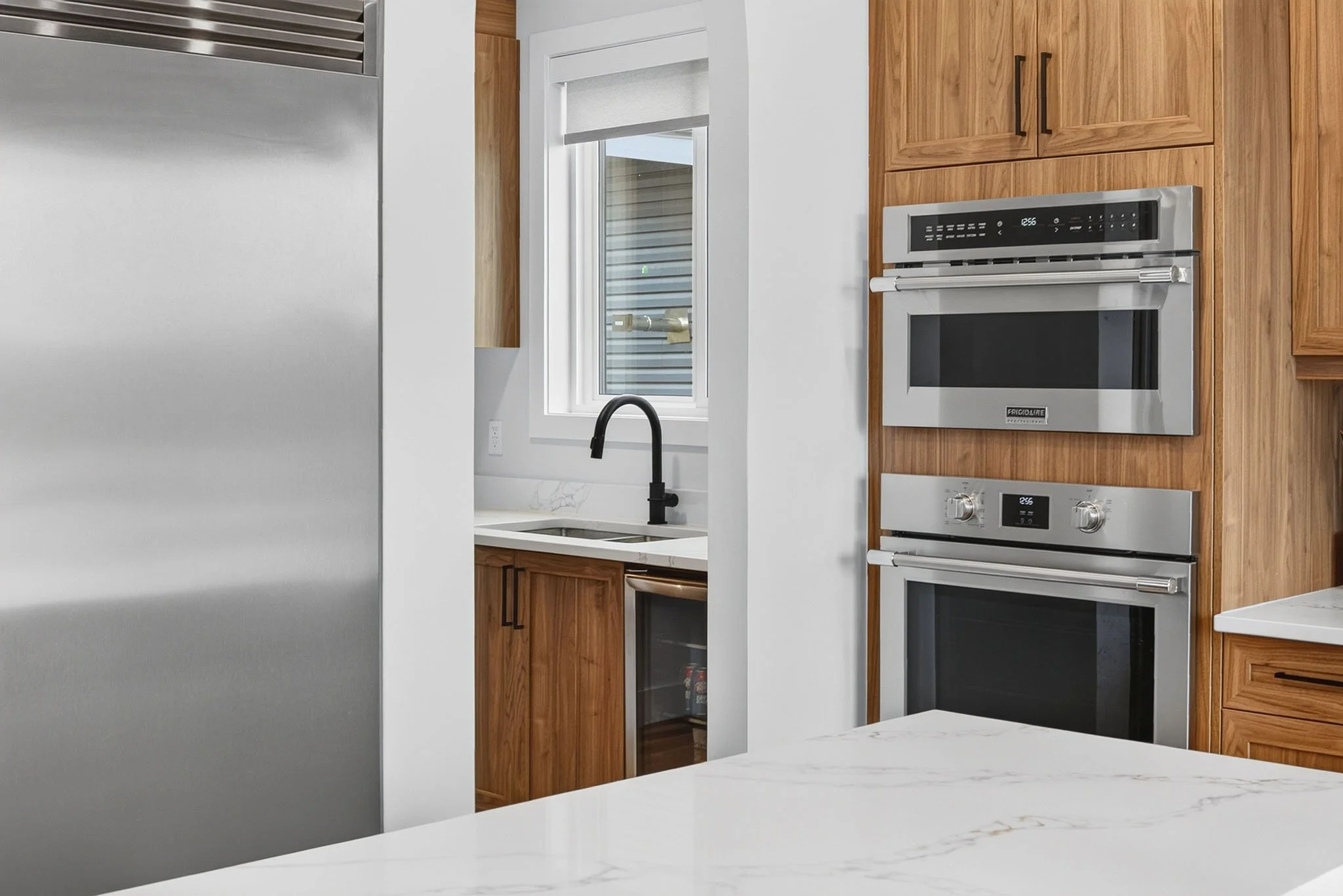 A modern kitchen with wooden cabinets, a built-in microwave and oven, a black faucet, a window with a roller shade, and a white marble countertop.