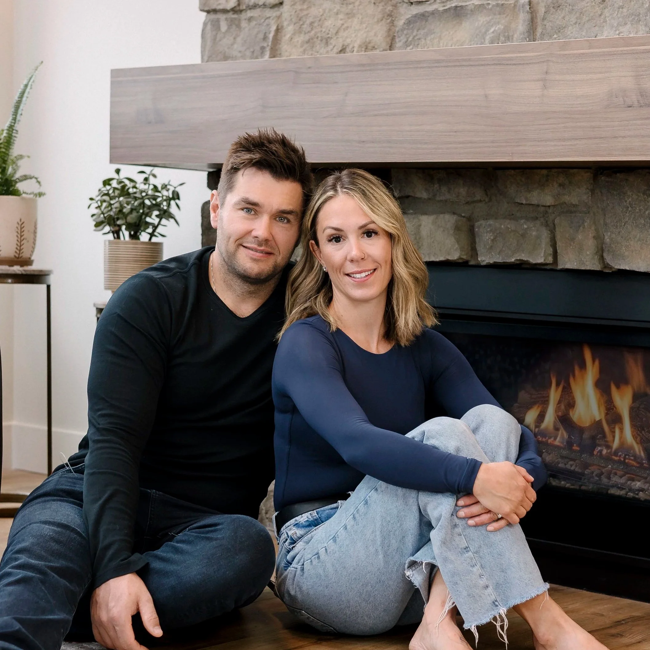 A man and woman sitting close together on the floor in a cozy room, with a stone fireplace in the background. The man has short dark hair and is wearing a black shirt. The woman has blonde hair with waves and is wearing a blue long-sleeve top and light blue ripped jeans. They are smiling and looking at the camera.
