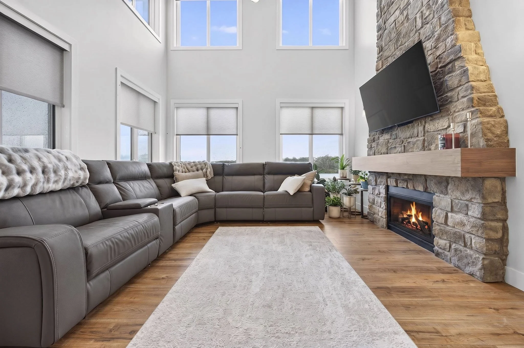 A modern living room featuring a large gray sectional sofa adorned with plush pillows, a white area rug, and a fireplace with a stone surround and a mounted flat-screen TV above it. The room has high ceilings and multiple windows allowing natural lig