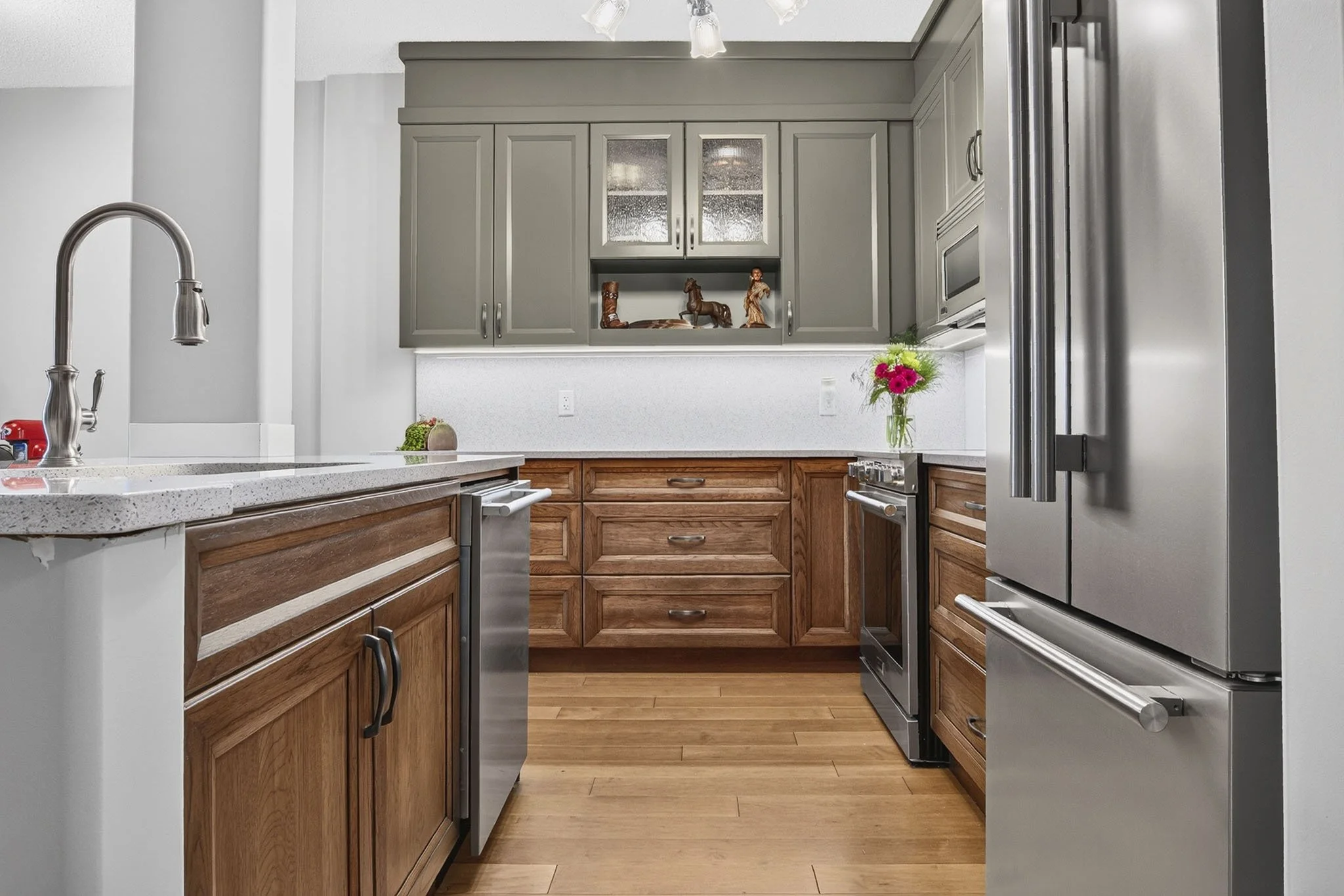 View of a kitchen with gray upper cabinets, wooden lower cabinets, a stainless steel refrigerator, and a marble countertop with decor and a vase of flowers.