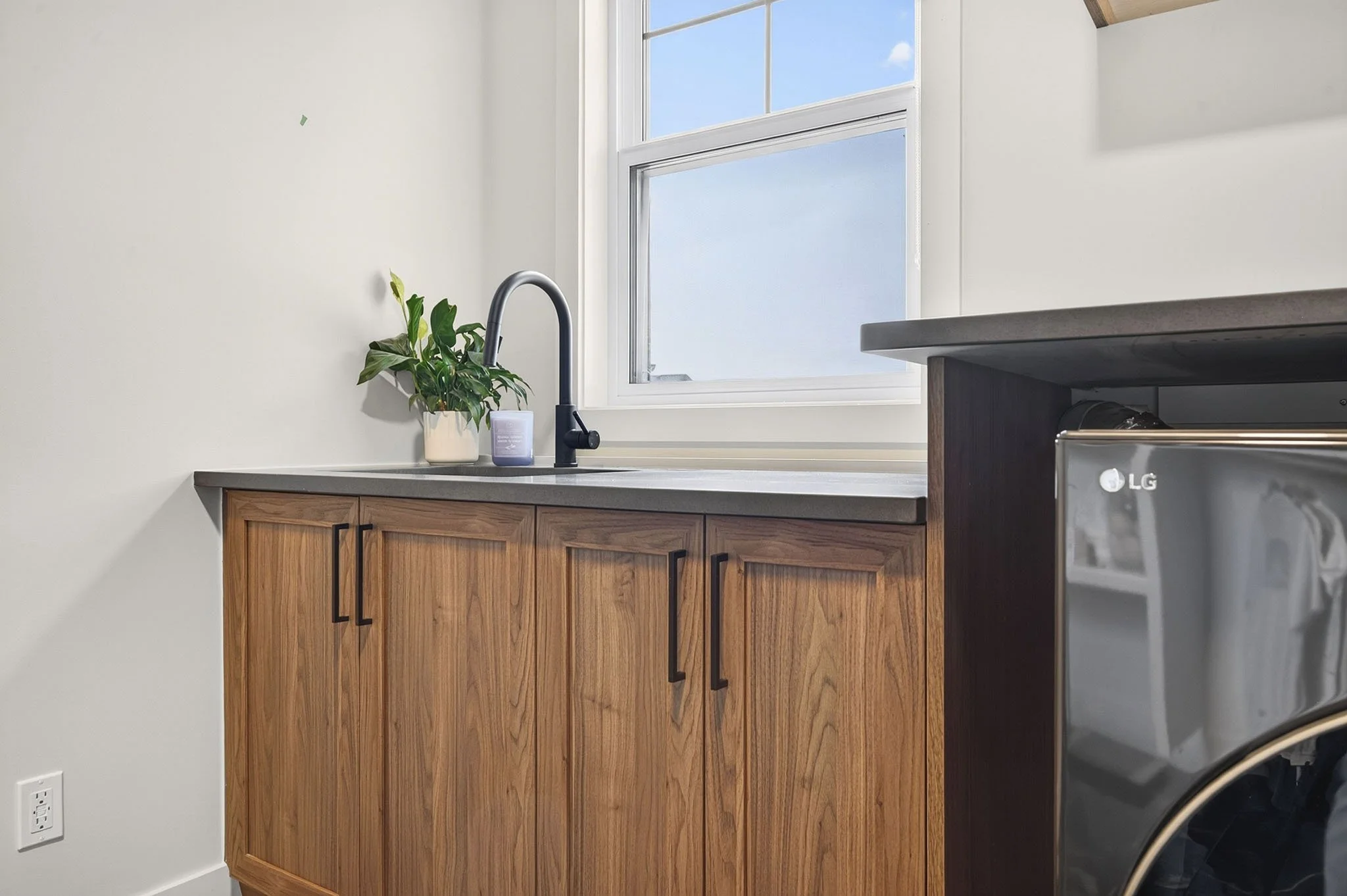 A kitchen corner with wooden cabinets, a black kitchen faucet, a potted plant, a soap dispenser, a window showing blue sky with clouds, and part of a black LG refrigerator.