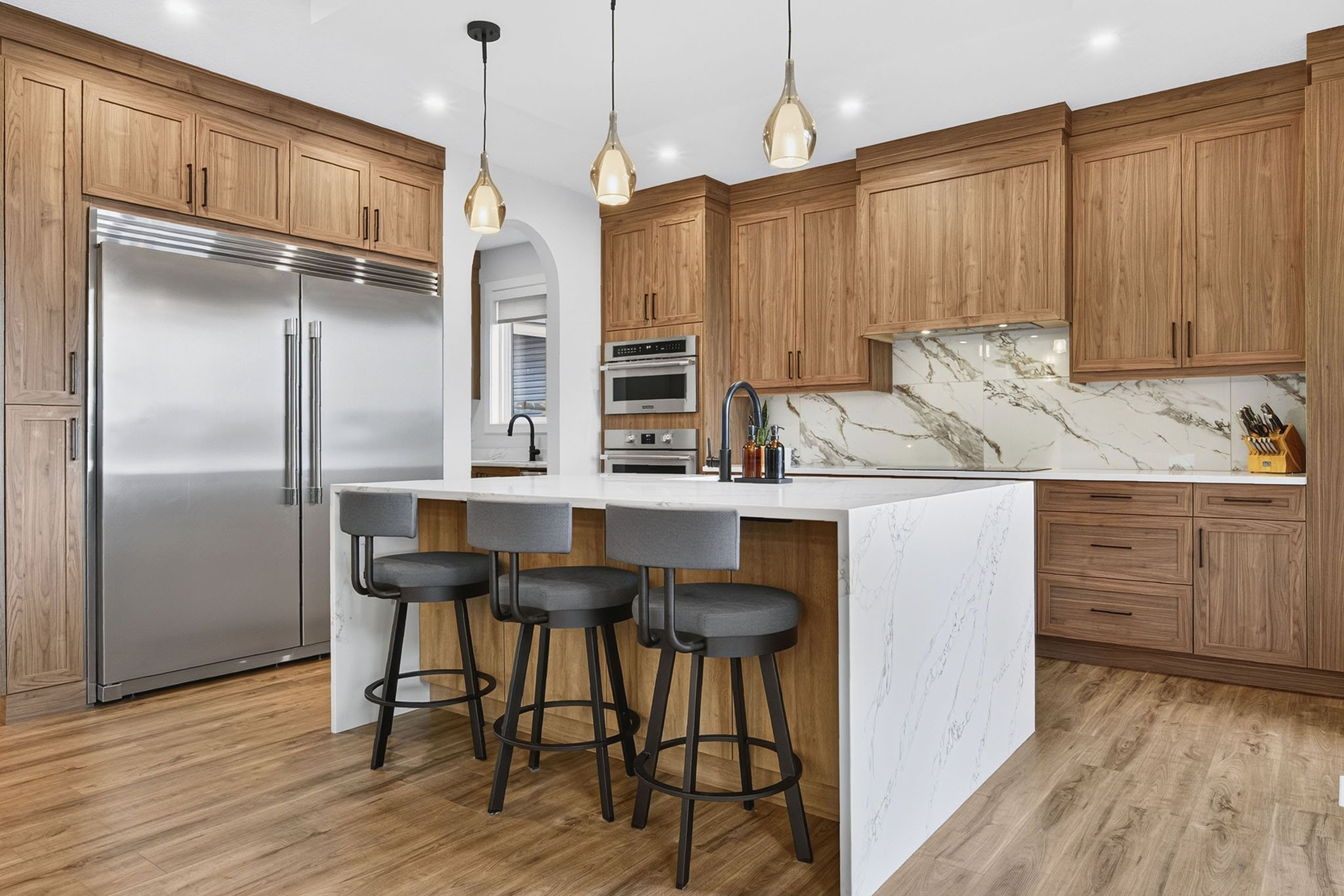 Modern kitchen with wooden cabinets, stainless steel refrigerator, marble backsplash, white island with bar stools, pendant lights, and wooden flooring.