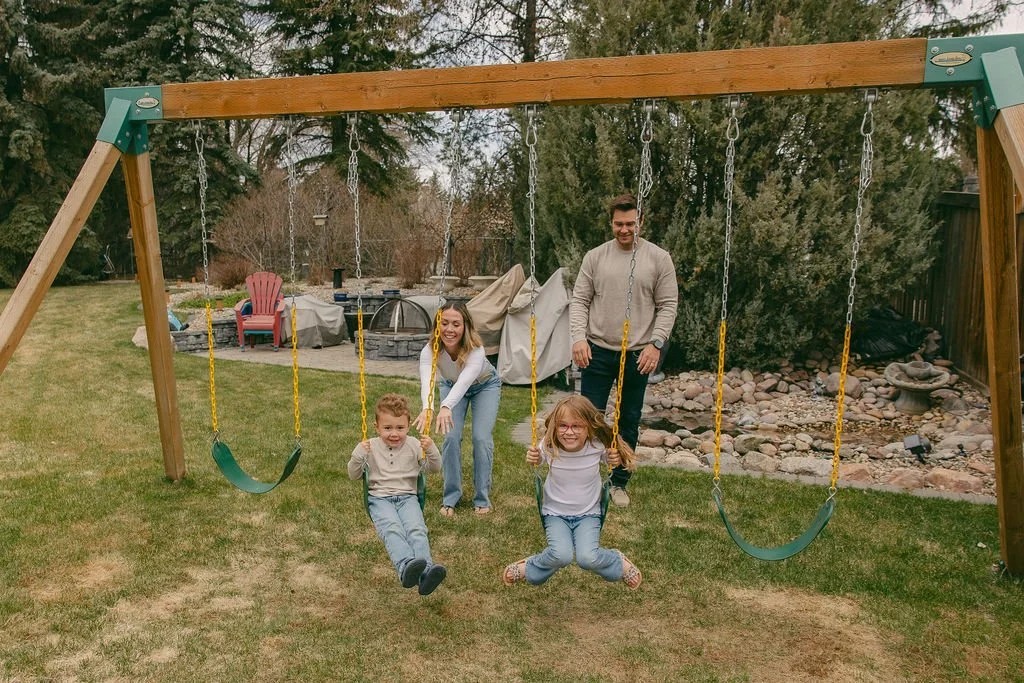 The Fosters - a family-owned homebuilding company enjoying time on a playground swing set in a backyard, with two children on swings and two adults standing behind them.