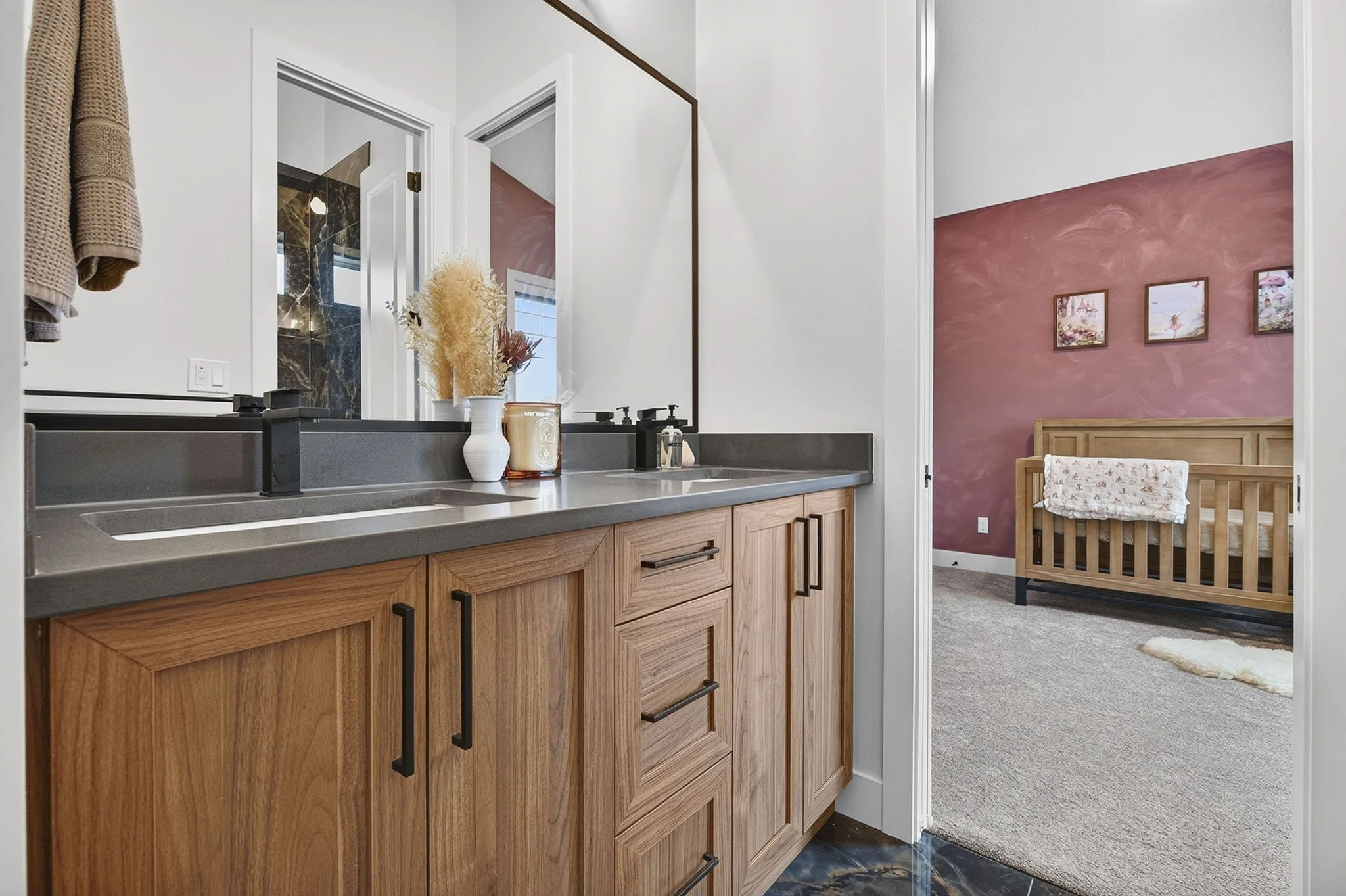 A view of a modern bathroom with a wooden vanity, black hardware, and decorative items on the countertop, overlooking a bedroom with pink textured wall, framed art, and a wooden bed frame with floral bedding.