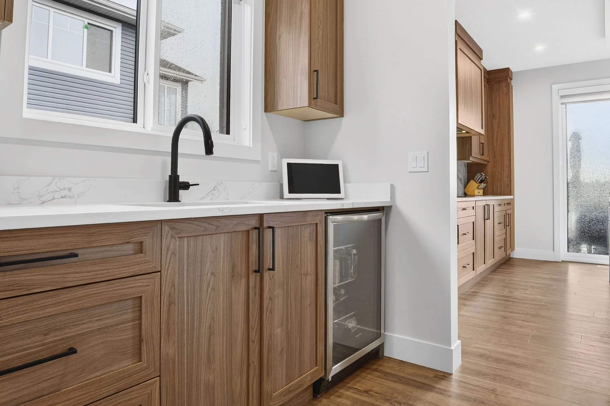 Kitchen with wooden cabinets, white marble countertops, a black kitchen faucet, a small white tablet, and a mini fridge, with a window showing a rainy day outside.