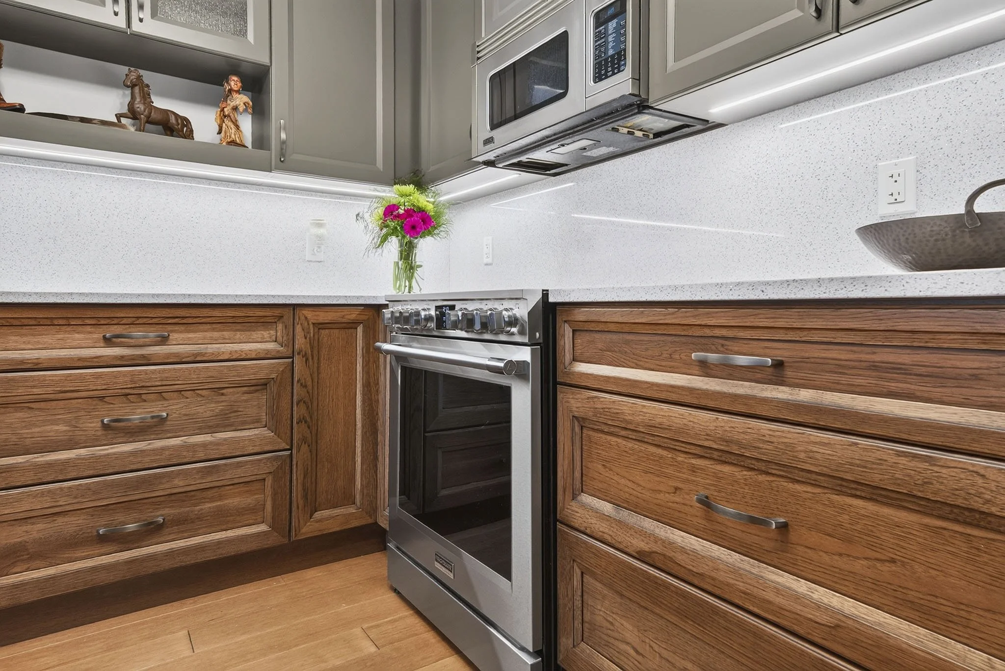 Kitchen with wooden cabinets, a white speckled countertop, a black oven with a silver handle, a stove, a microwave above, an electrical outlet, a brown pot, a vase with pink and green flowers, and decorative statues on the shelf.