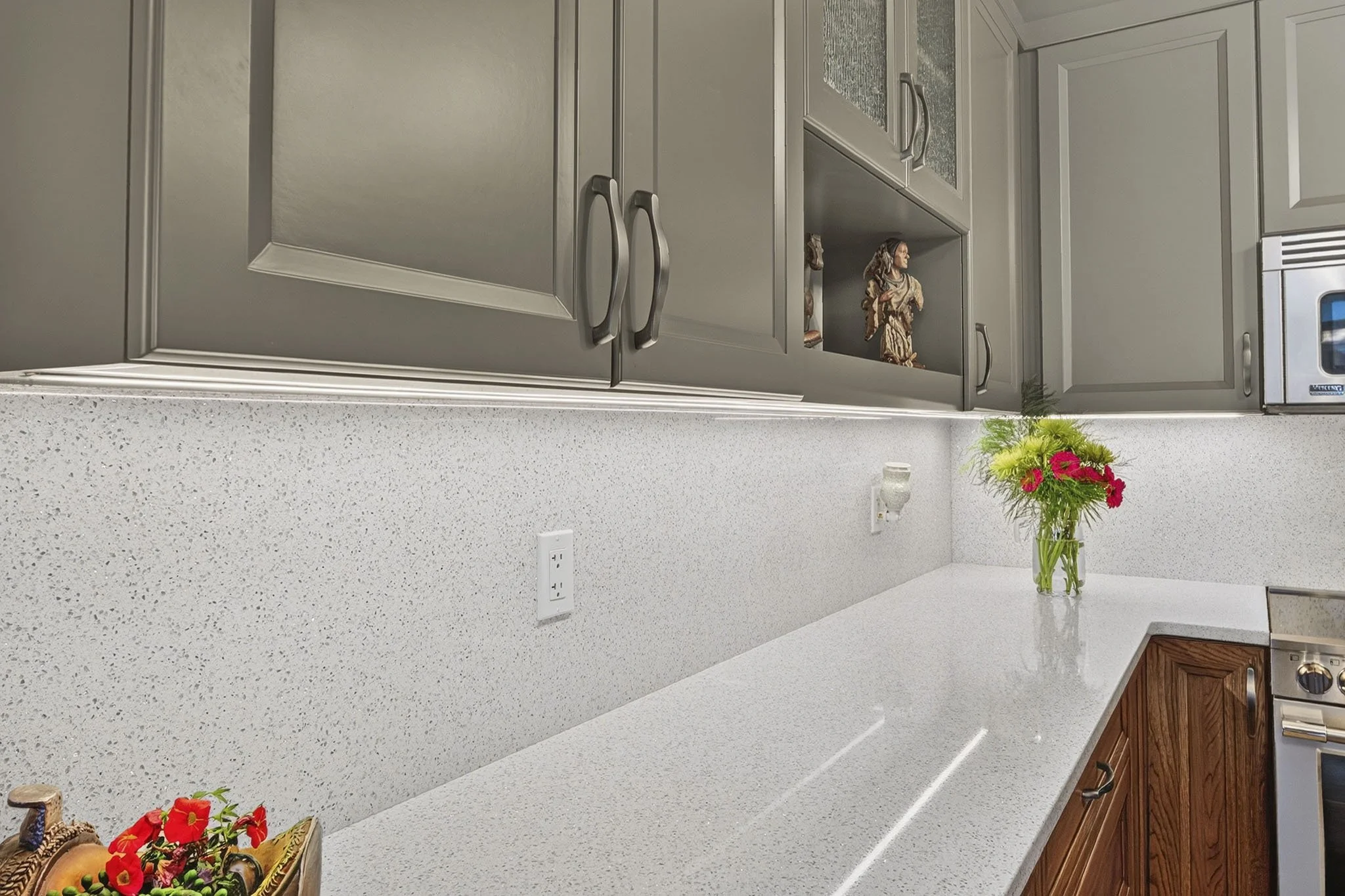 A kitchen with gray cabinets, white speckled countertop, a small clay angel figurine, a vase with pink flowers, and a window with blinds.