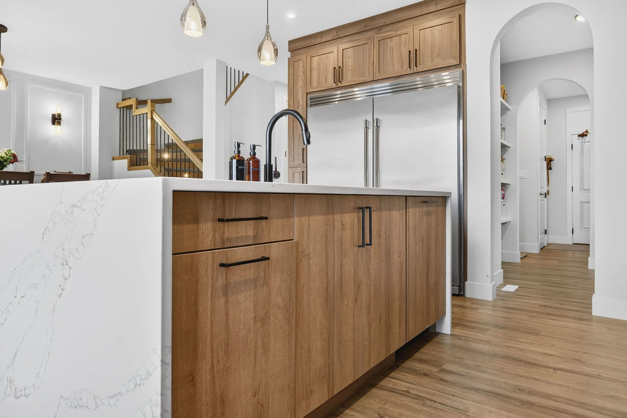 Modern kitchen with wooden cabinets, stainless steel refrigerator, white marble Island with black handles, black faucet, and pendant lights, hardwood floors, open archway.