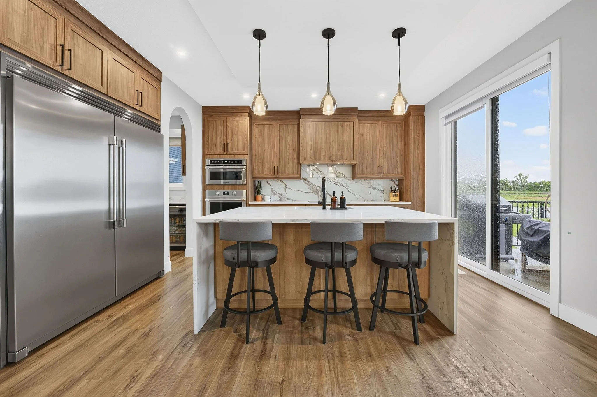 Modern kitchen with wooden cabinets, stainless steel refrigerator, marble backsplash, island with four gray bar stools, large sliding glass door showing outdoor patio and greenery, pendant lights above island, hardwood floors.