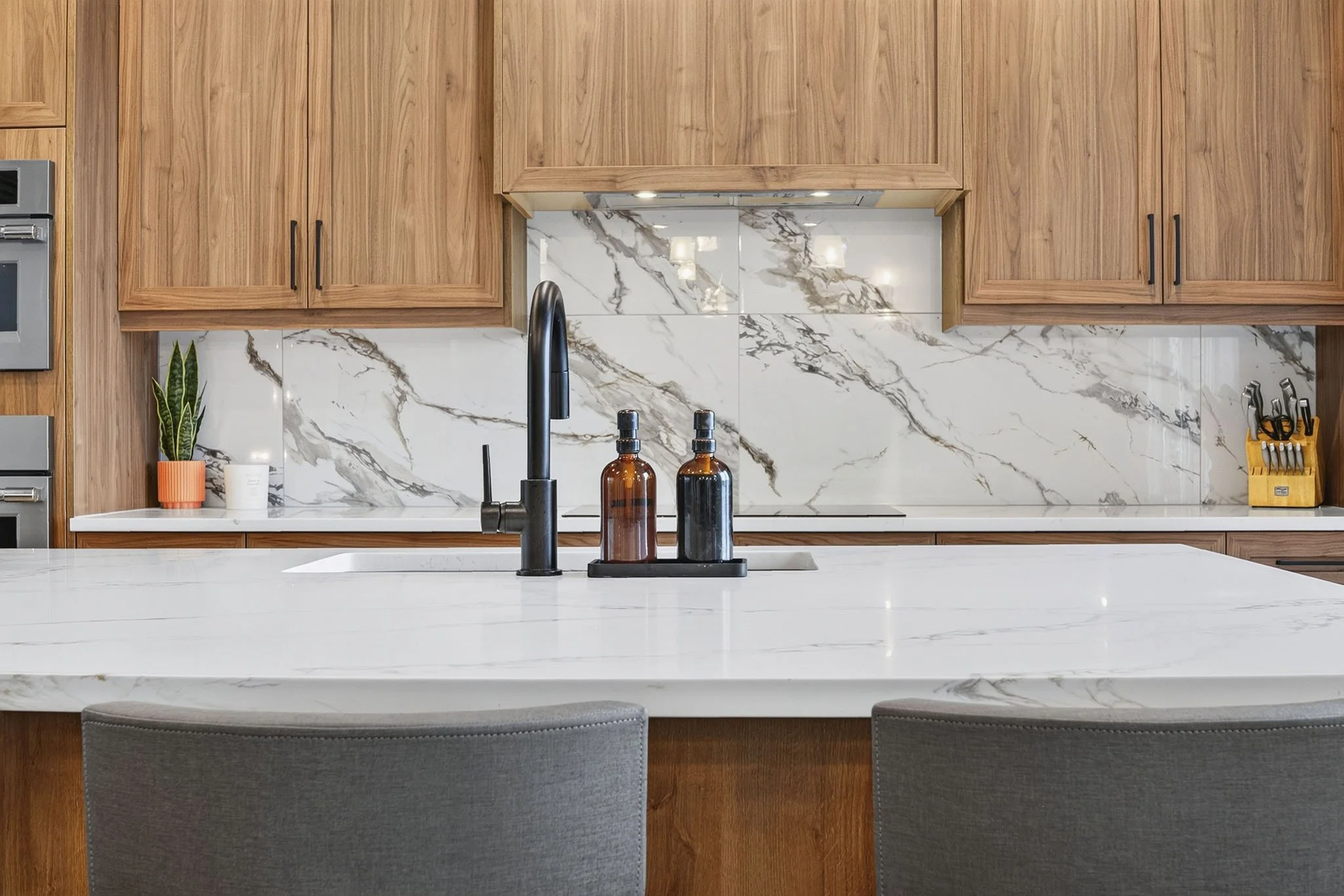 Kitchen with light wood cabinets, marble backsplash, and a white countertop with a black faucet. There are two amber bottles, a small plant in an orange pot, and other small items on the countertop. Part of a gray stool is visible at the bottom of th