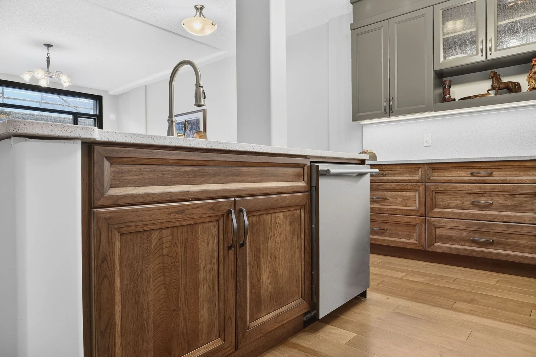 Kitchen interior with wooden cabinets, a granite countertop, a stainless steel dishwasher, and a street view through a large window.