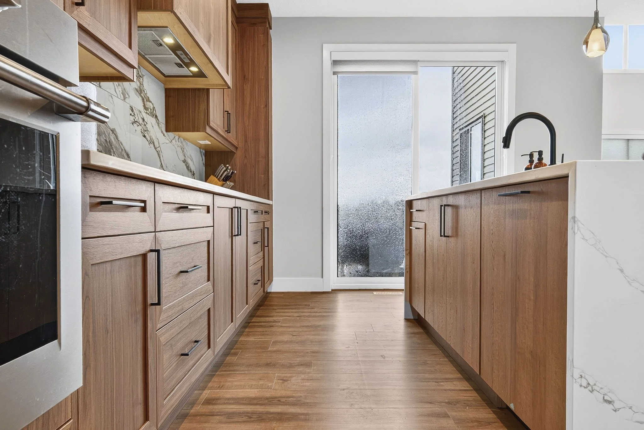 A modern kitchen with wooden cabinets, a marble backsplash, and a glass door leading outside. The kitchen features a black faucet, a white countertop, and a few bottles of soap or lotion on the sink. There is a window above the sink letting in natura