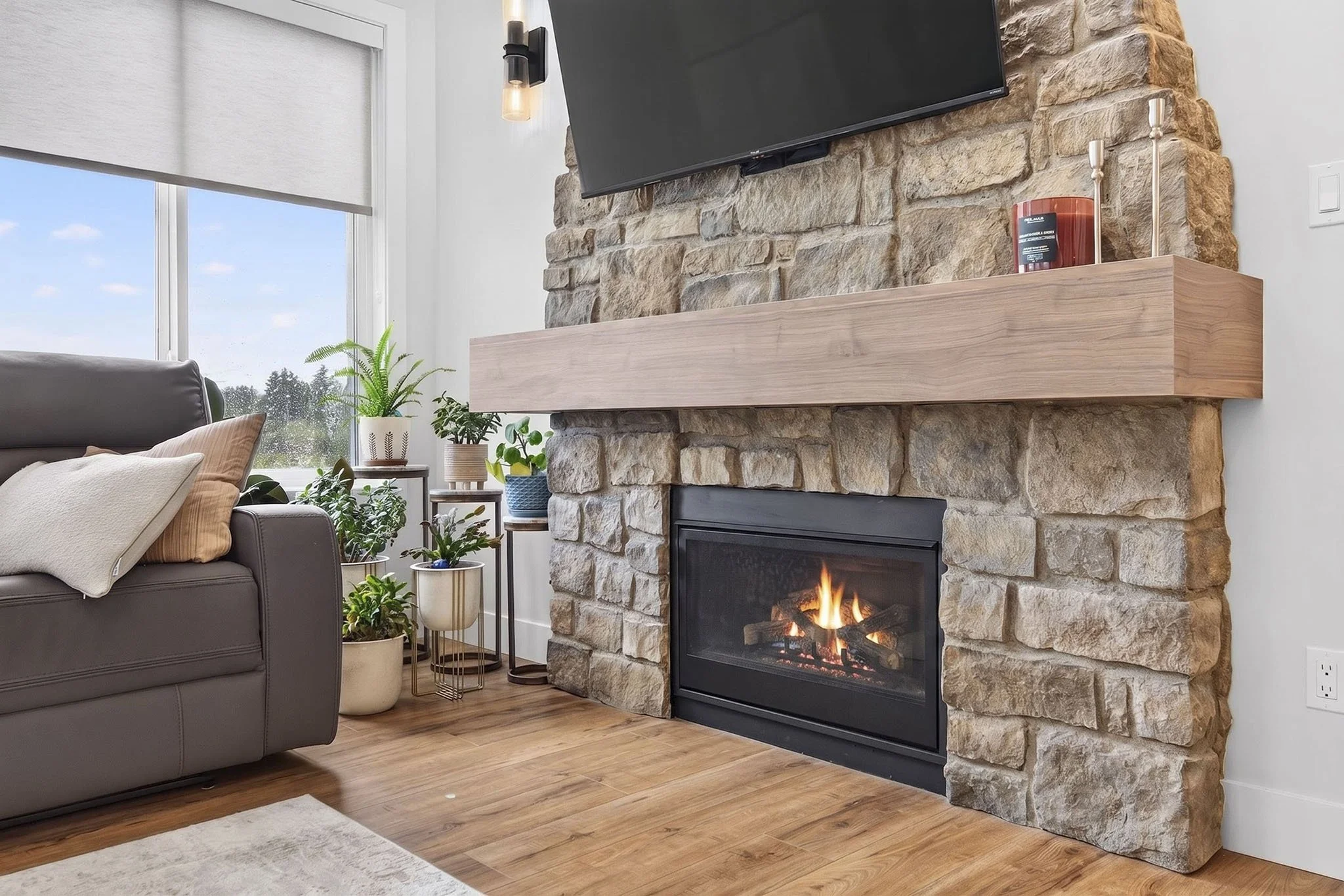Living room with a fireplace built from large stones, a flat-screen TV mounted above it, and wooden mantel; cozy seating area with pillows, a gray armchair, and various potted plants near a window with a white blind; hardwood flooring.
