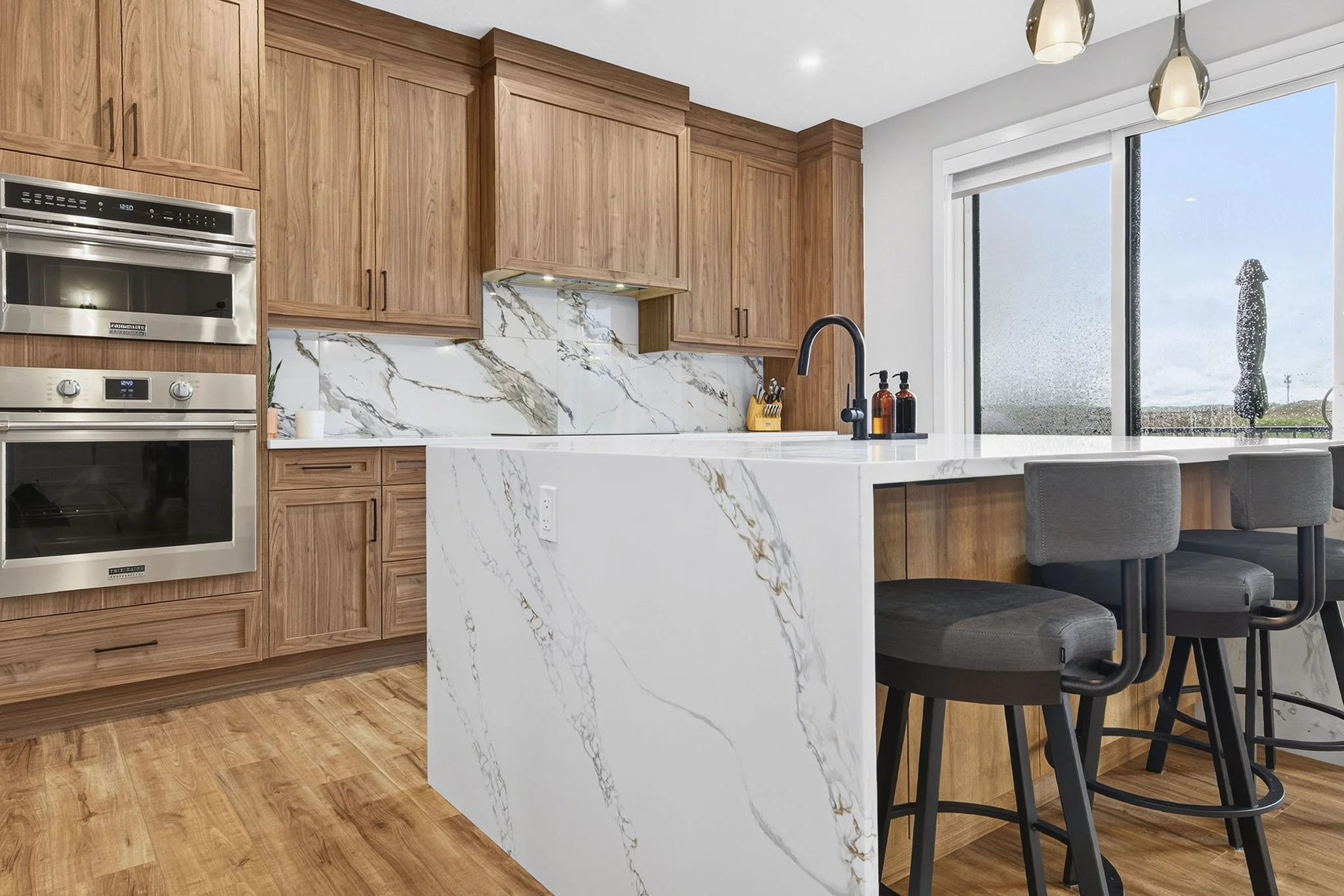 Modern kitchen with wooden cabinets, a marble island, and a large window with a rural view.