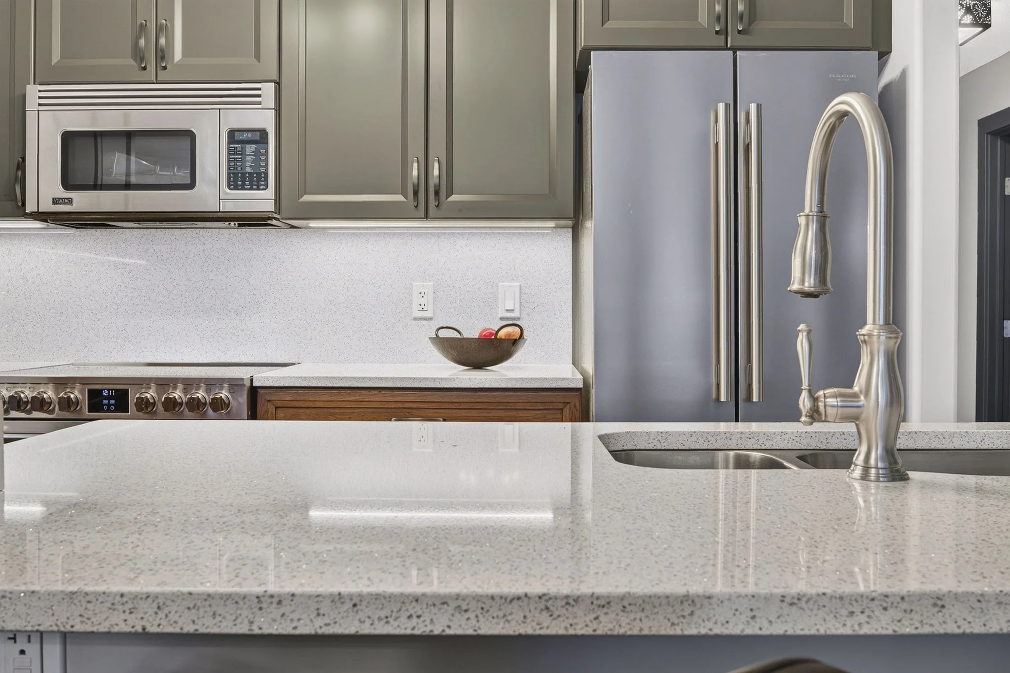 Kitchen with gray cabinets, stainless steel microwave and refrigerator, white speckled countertops, a black bowl with oranges, and a modern silver faucet.