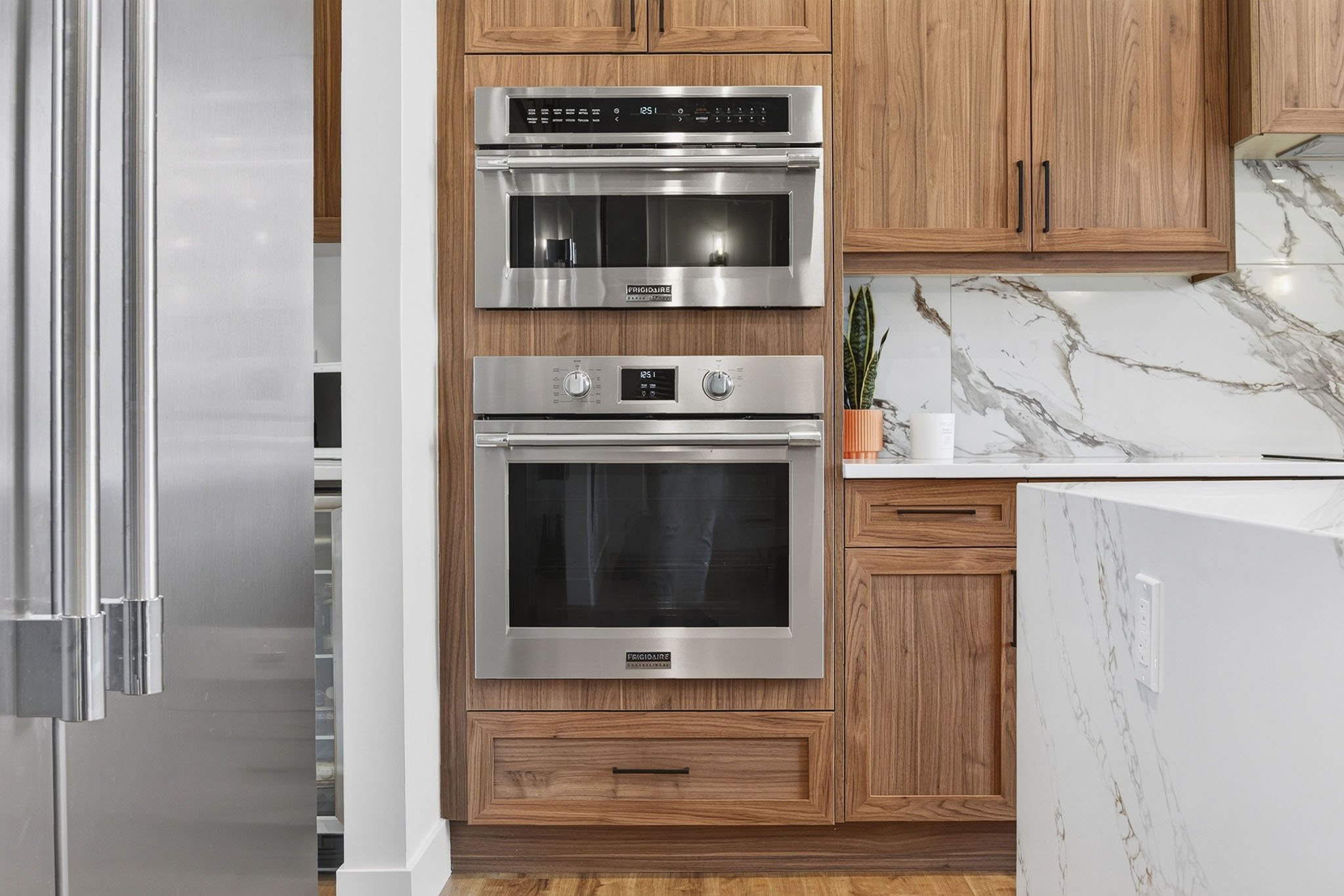 Modern kitchen with wooden cabinets, stainless steel double wall oven, marble countertop, part of a refrigerator.