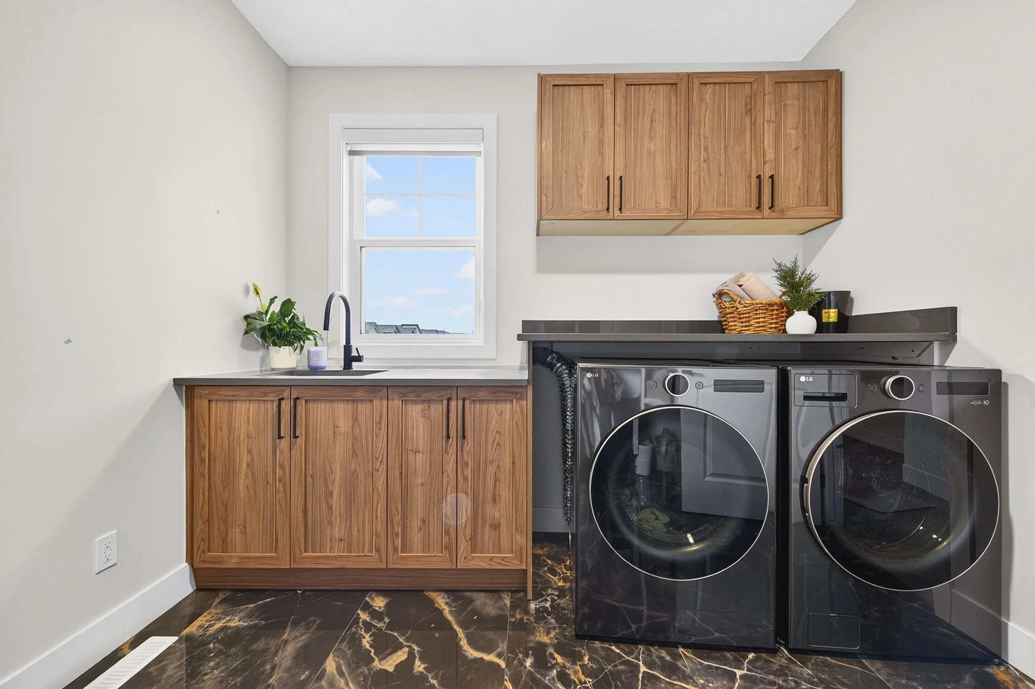 A laundry room with white walls, a window with a view of blue sky and clouds, wooden cabinets, a plant on the counter, and a black washing machine and dryer with marble-look flooring.