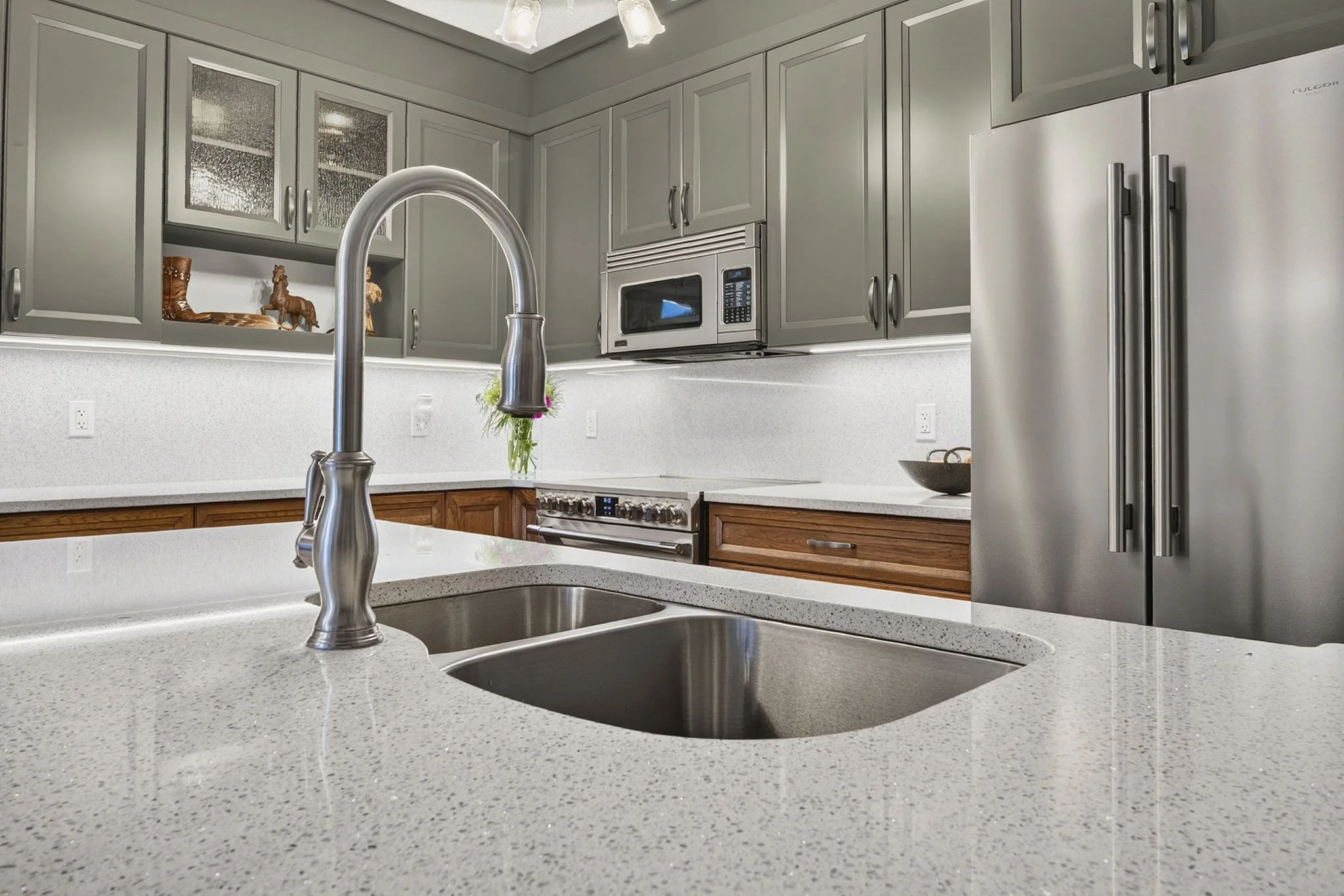 A modern kitchen with white speckled countertops, gray cabinets, a stainless steel refrigerator, built-in microwave, and a stainless steel sink with a tall curved faucet. Decorative items, such as figurines and a bowl, are visible on the shelves. Pen