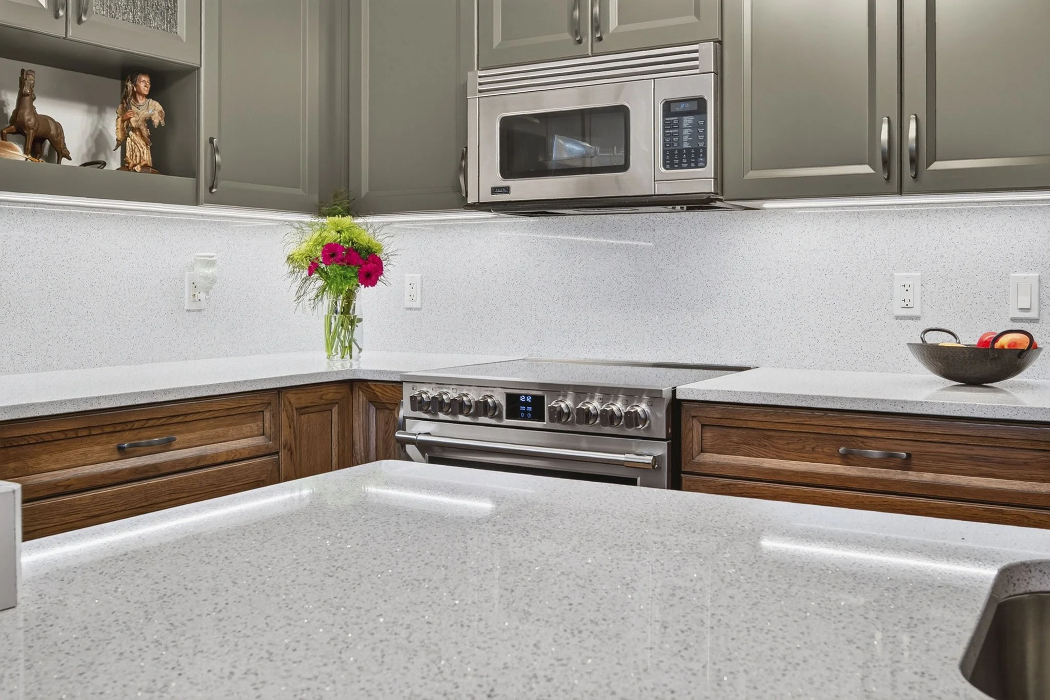 A kitchen with light gray walls, gray upper cabinets, and dark wood lower cabinets. There is a stainless steel microwave above a matching oven, a vase of pink flowers on the countertop, and decorative figures on an open shelf. There are electrical ou
