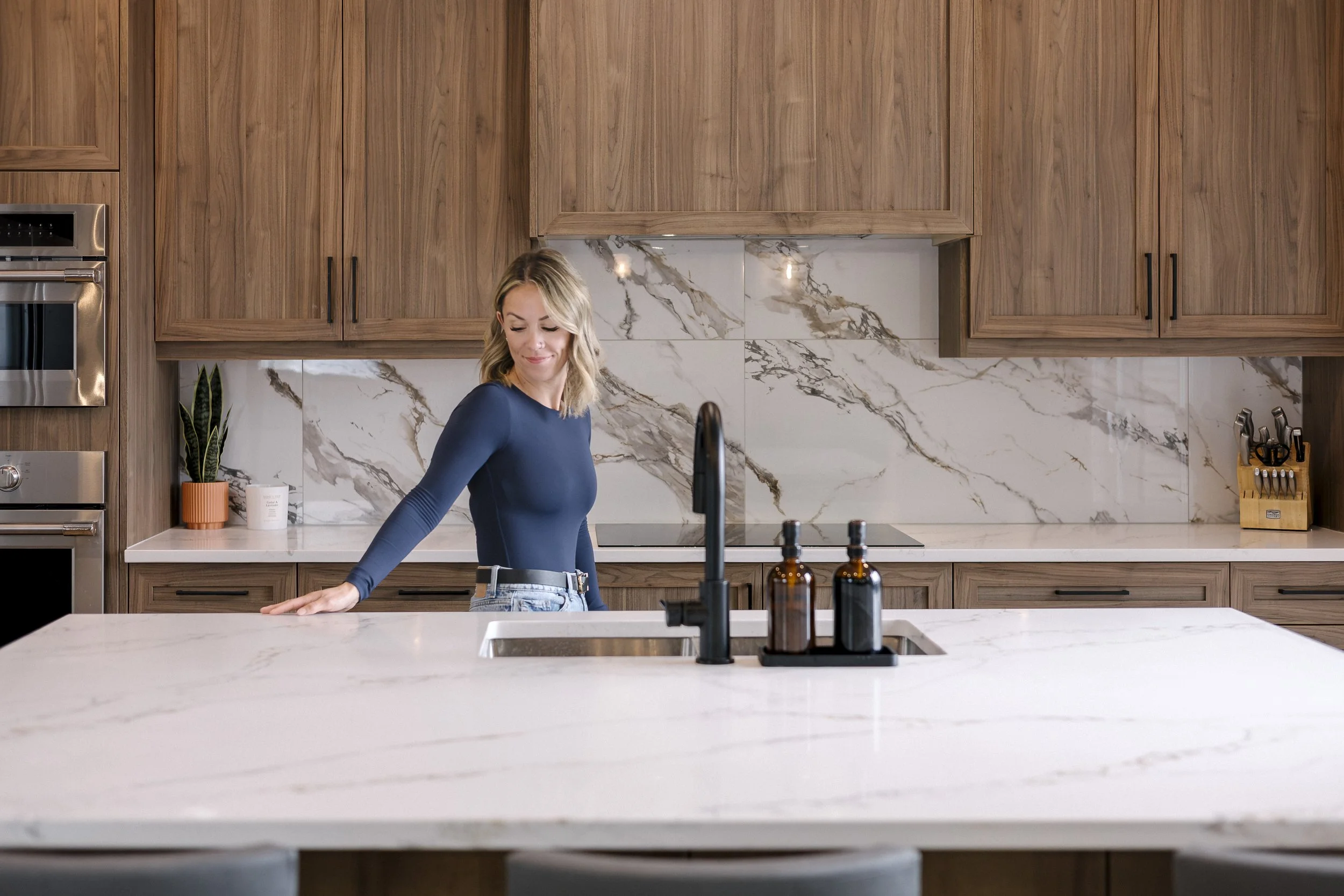 A woman with blonde hair wearing a blue long-sleeved top and jeans, standing in a modern kitchen with wooden cabinets, marble backsplash, and a white marble countertop. There are two brown bottles near the sink and a potted plant on the counter.
