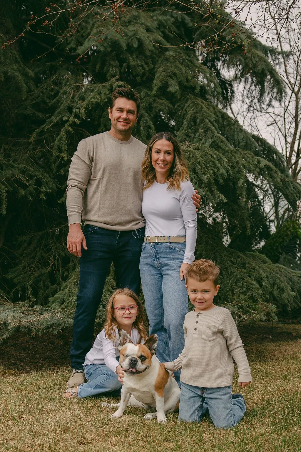 Chelsea and Jeff Foster pose with their children for CJ Living Co. The parents stand behind the children, who are sitting or kneeling on the grass, with the dog in front. The family is smiling and dressed casually.