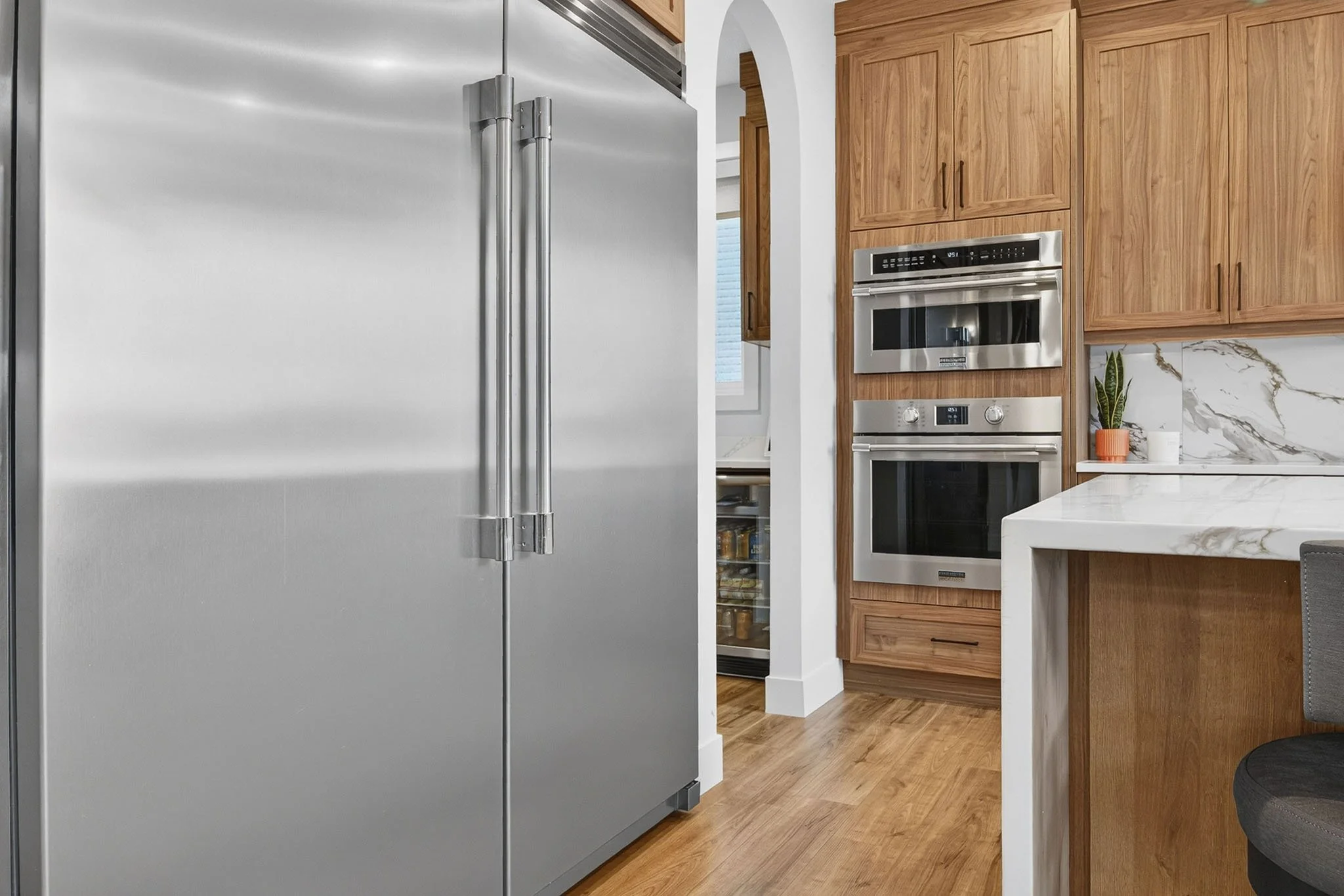 Kitchen with a large stainless steel refrigerator on the left, wooden cabinets, built-in microwave and oven, marble countertop, and a plant in an orange pot.