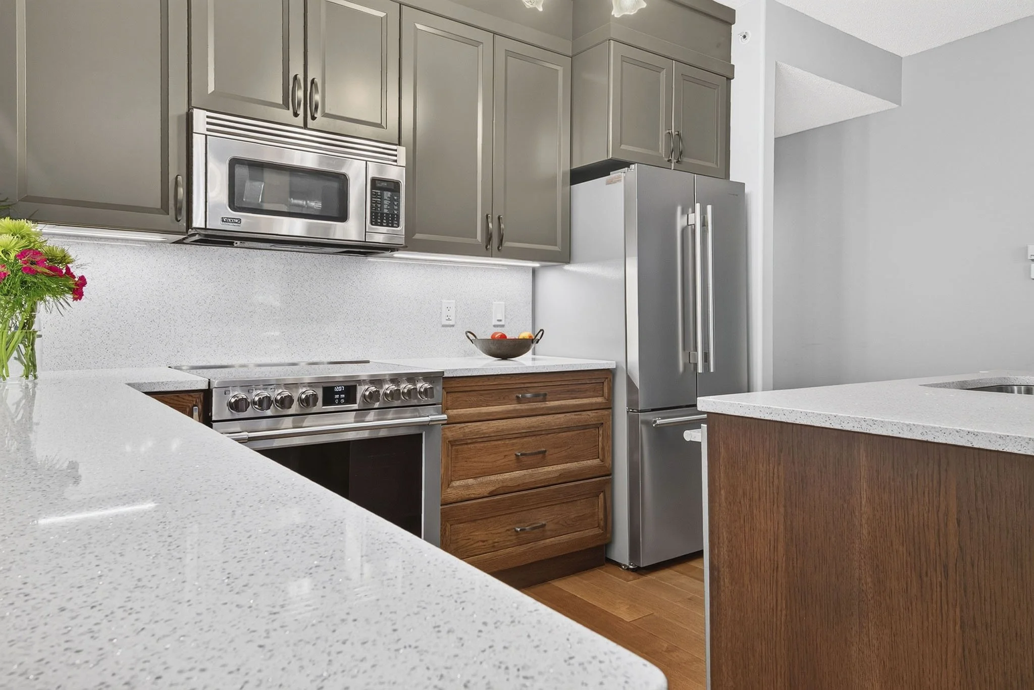 Modern kitchen with light gray cabinets, stainless steel microwave and refrigerator, white speckled countertops, and a wooden drawer cabinet. A black bowl with apples and a bunch of pink flowers are on the counter.
