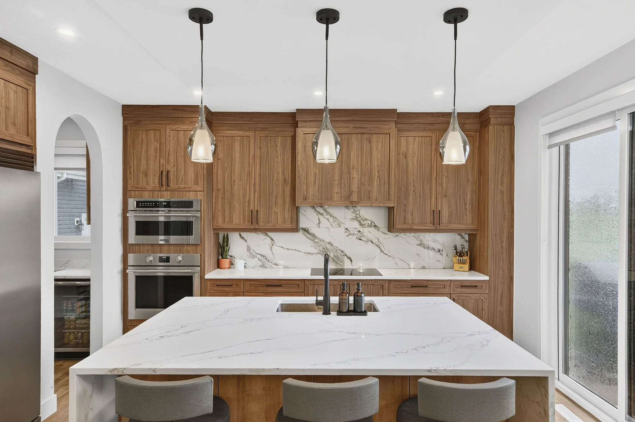 Modern kitchen with wooden cabinets, white marble backsplash, and a large white marble island with a sink. Three pendant lights hang above the island. There is a window on the right side and a built-in oven and microwave on the left.