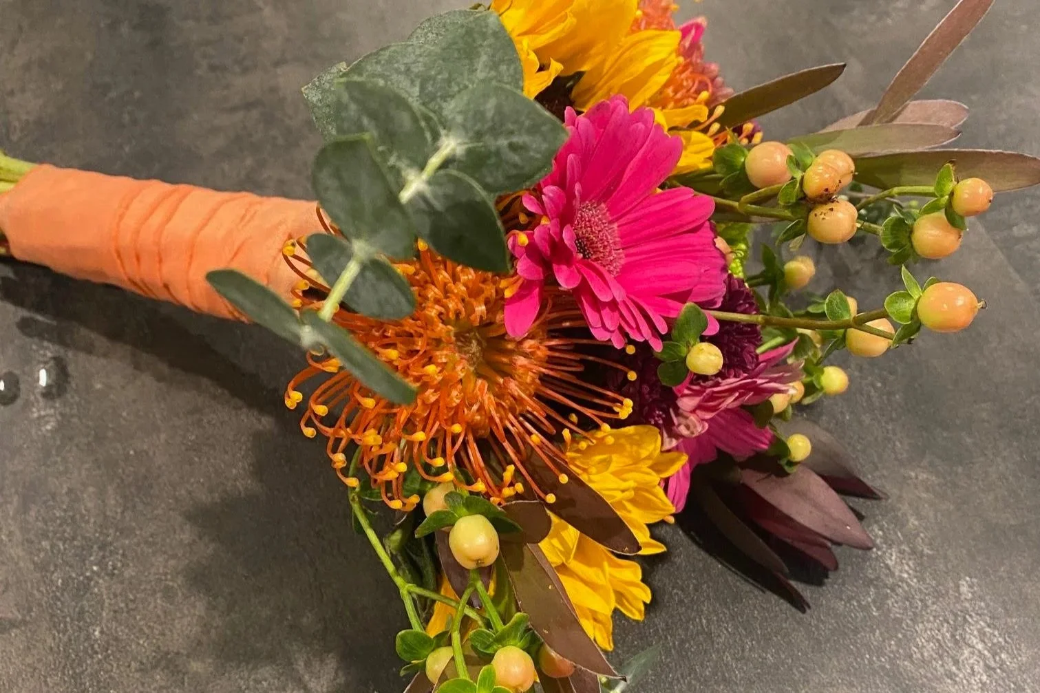 A colorful boutonniere with pink, yellow, and orange flowers, green leaves, and an orange carrot on the left side, resting on a dark surface.