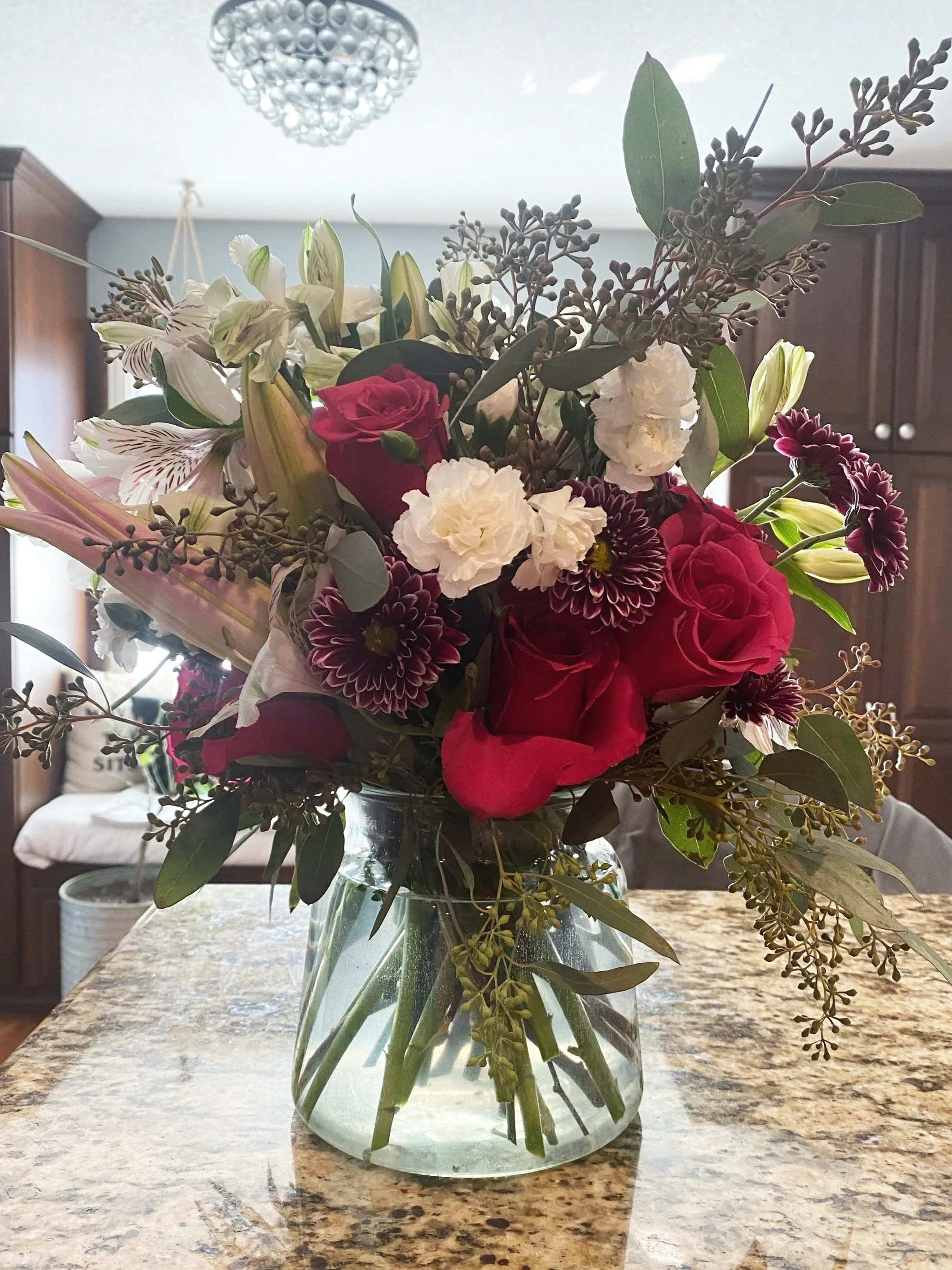A mixed bouquet of flowers in a glass vase on a granite countertop, with a chandelier above and wooden cabinets in the background.