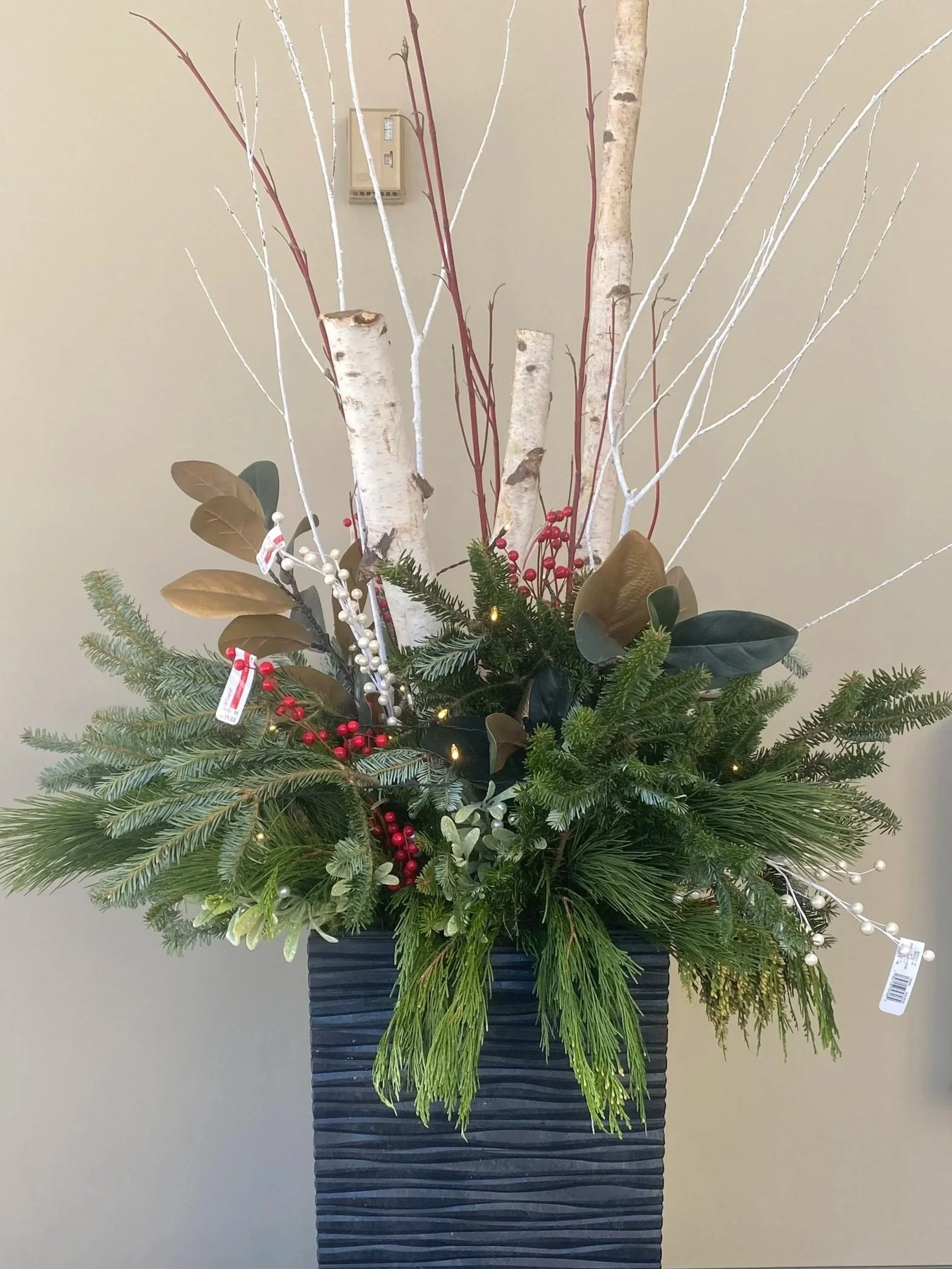 Holiday floral arrangement with white birch branches, evergreen branches, red berries, and white decorative elements in a tall black vase.