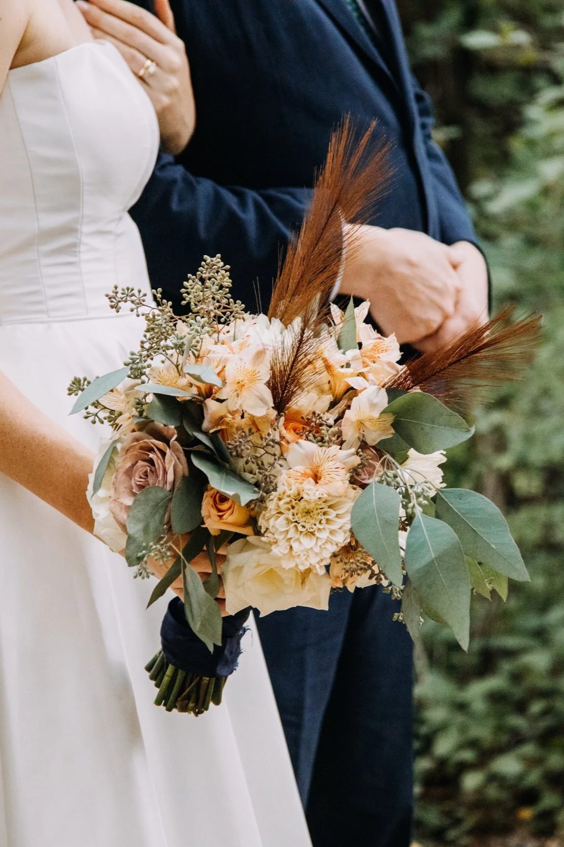 A bride holding a bouquet of peach and cream-colored flowers with green leaves and brown accents, beside a groom dressed in a dark suit. The couple's hands are intertwined, and the background shows greenery.