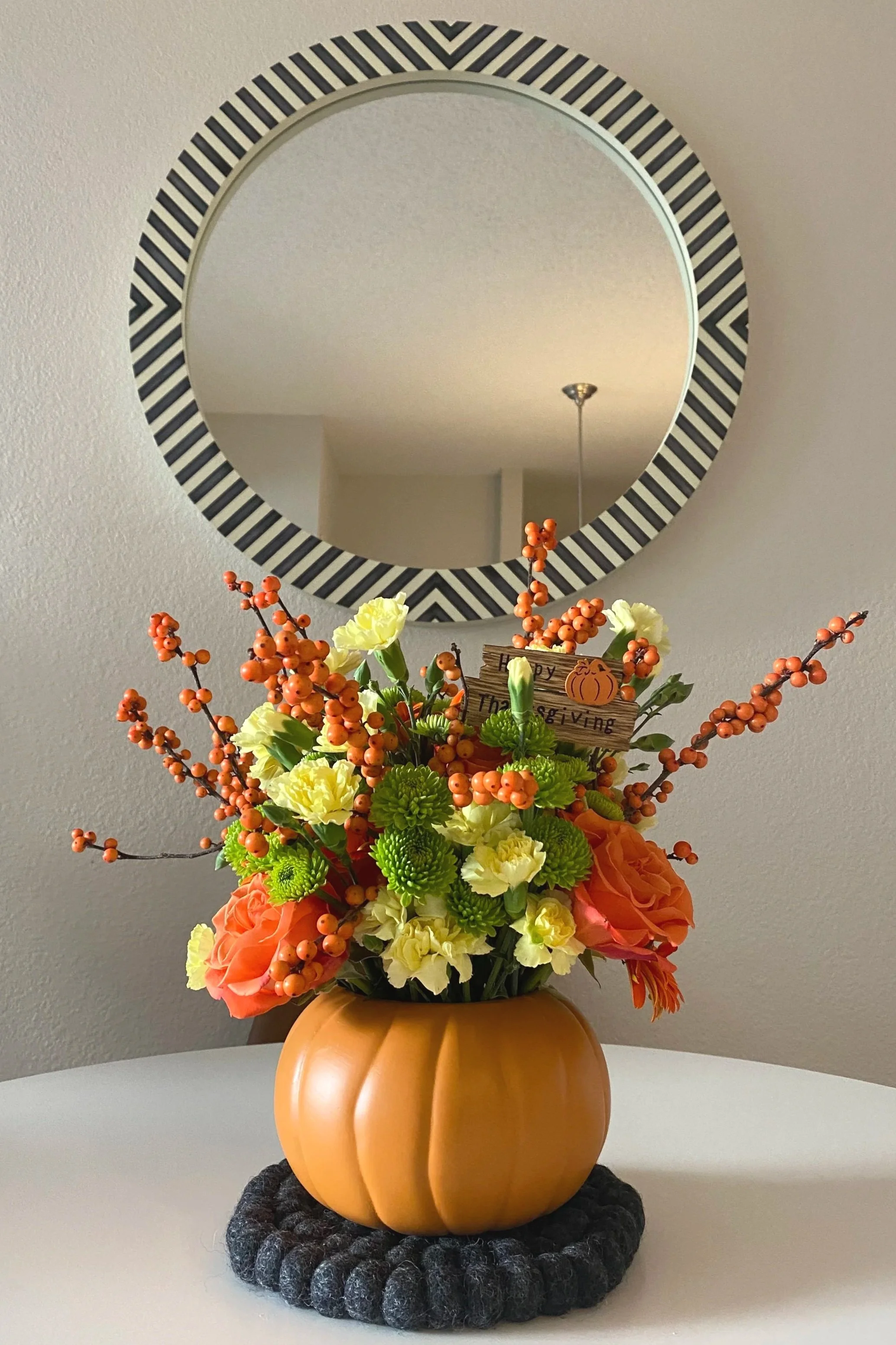A fall-themed flower arrangement with orange, yellow, and green flowers in a pumpkin-shaped vase, placed on a white table with a black felt mat underneath. A mirror with a black and white striped border hangs on the wall behind it.