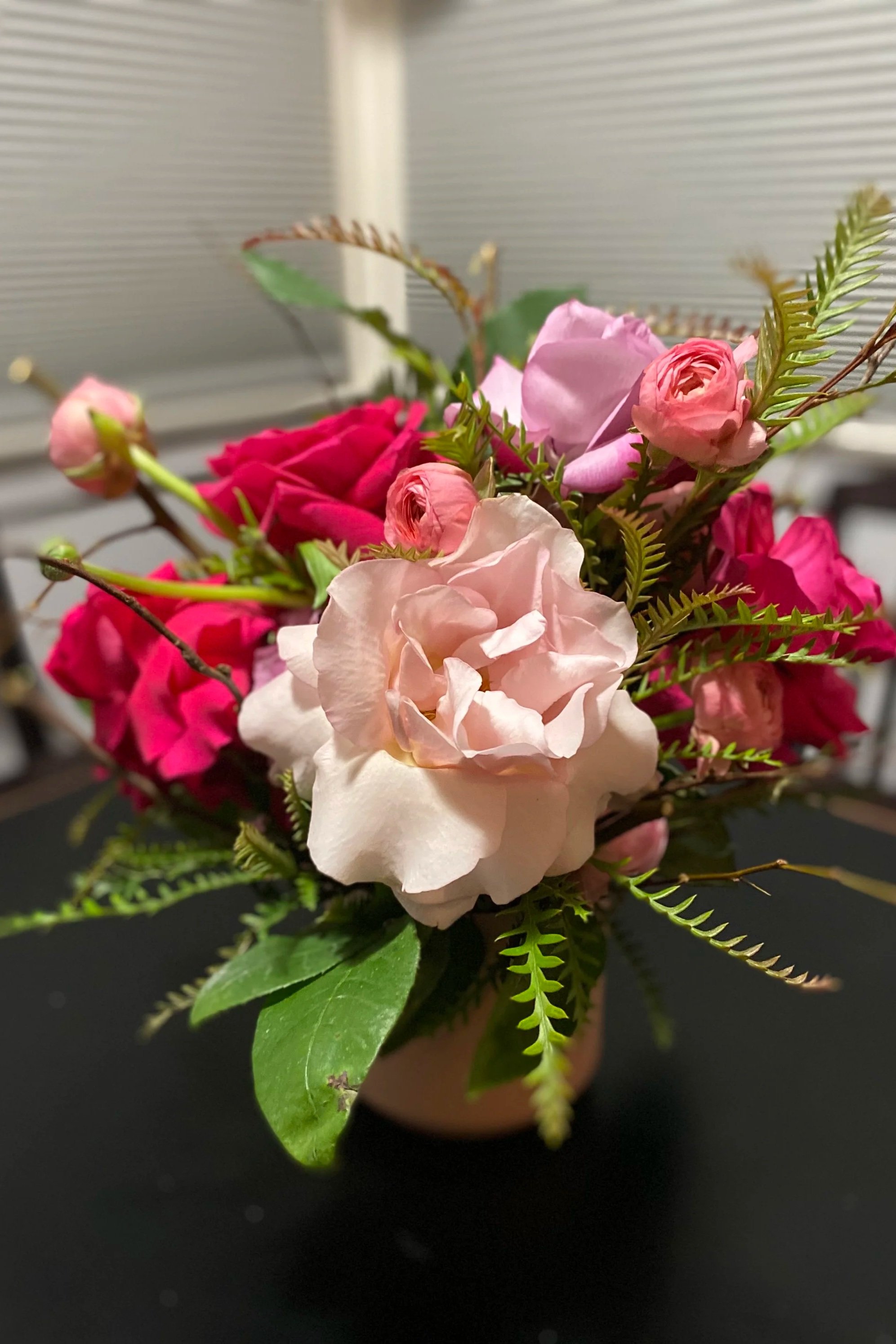 A small floral arrangement in a pink pot, featuring pink roses, light pink and dark pink spray roses, green fern-like leaves, and additional greenery, placed on a black surface with a blurred background.