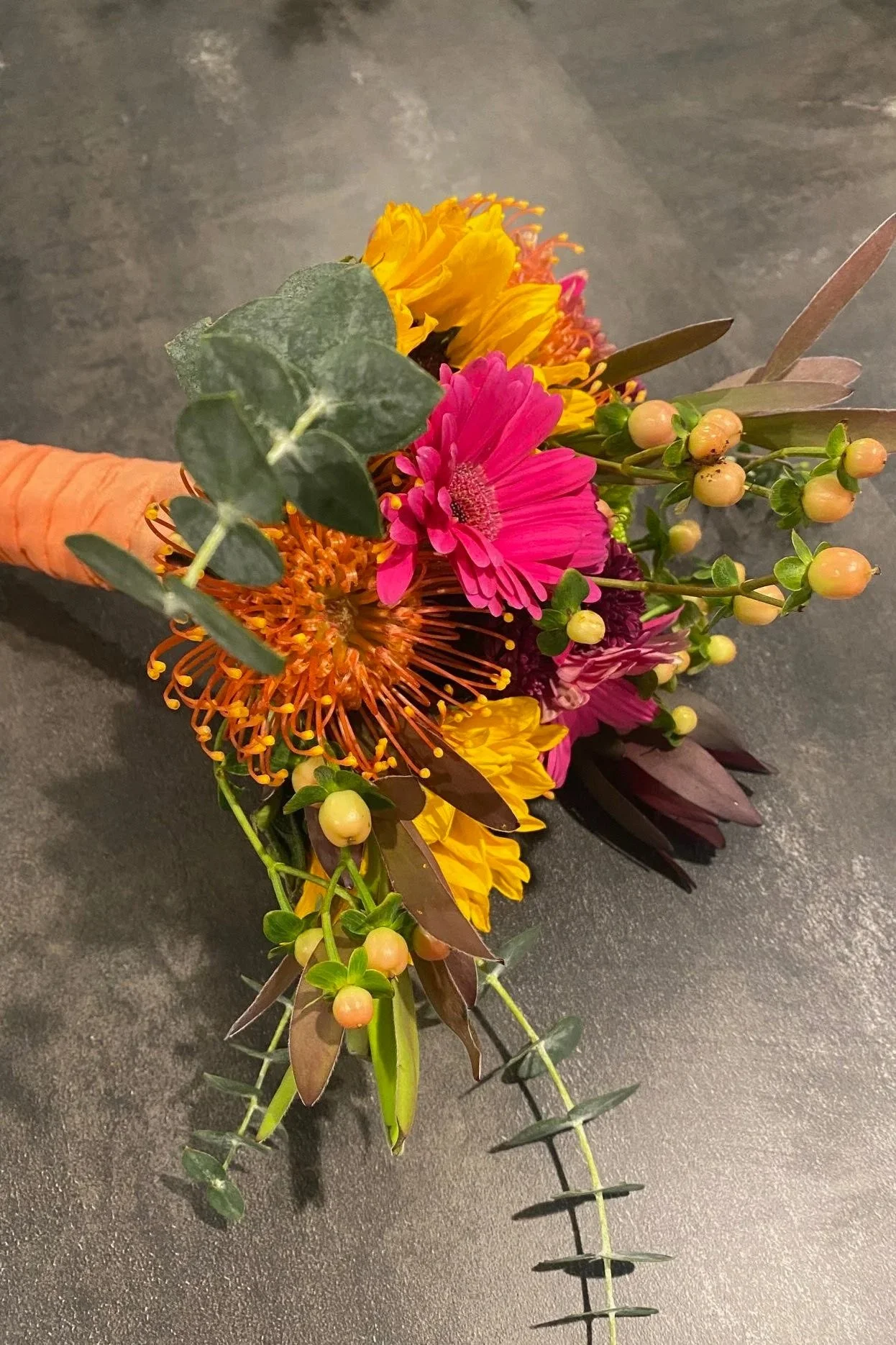 A vibrant bouquet with pink, yellow, orange, and purple flowers, including greenery and small berries, resting on a dark surface.