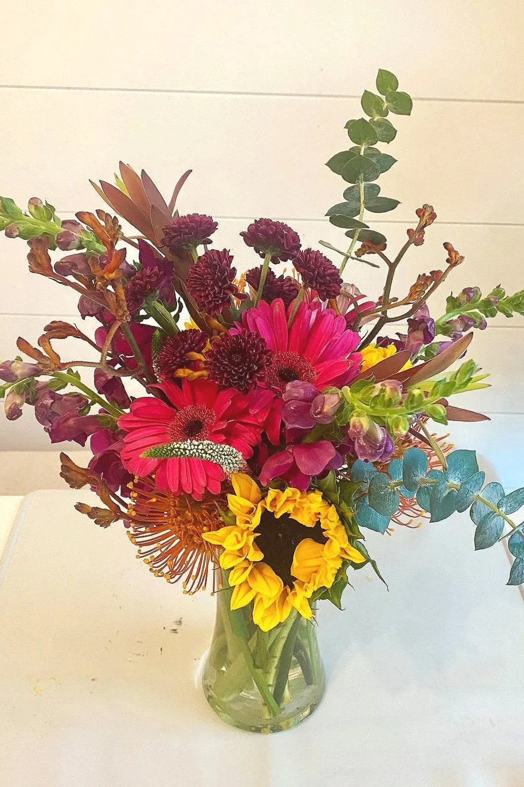 Colorful flower bouquet with pink, purple, yellow, and green flowers in a small glass vase on a white surface.