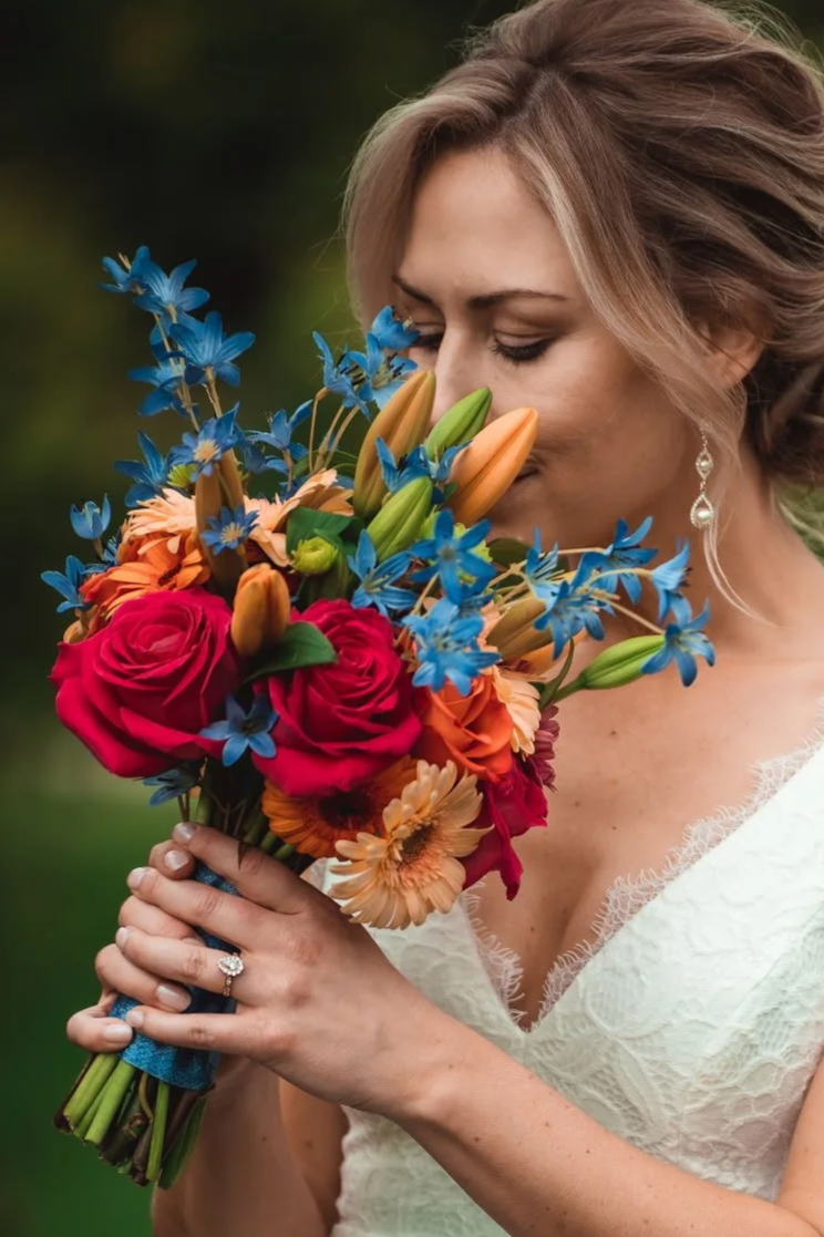 A woman in a white lace dress holding a colorful bouquet of flowers close to her face with her eyes closed.