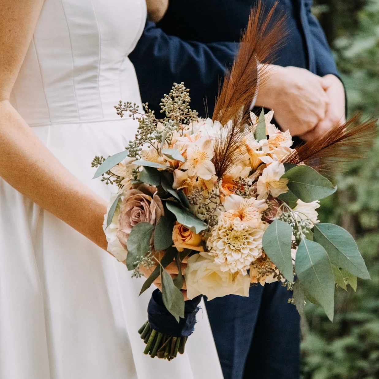 A bride holding a bouquet of cream and peach-colored flowers, greenery, and brown feathers, with a groom in a navy suit standing nearby.