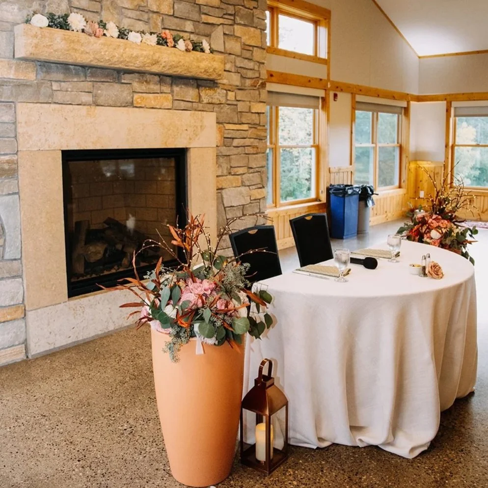 Indoor event setup with a round table decorated with floral arrangements, glasses of water, and small notepads, near a stone fireplace and multiple windows.