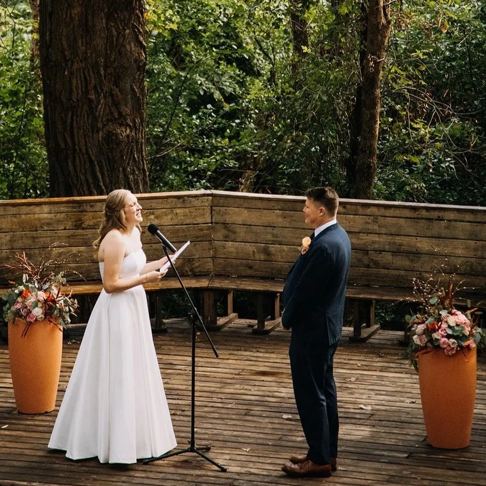 A wedding ceremony outdoors on a wooden deck with a woman in a white wedding dress and a man in a dark suit standing across from each other, exchanging vows, with floral arrangements on either side, dense greenery, and large trees in the background.