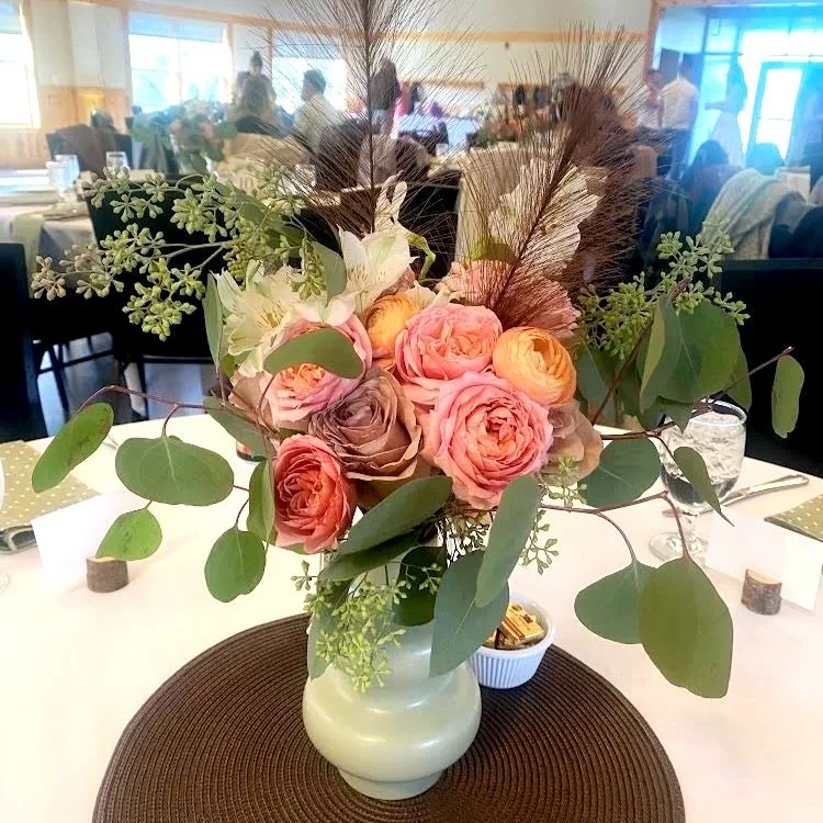 A floral centerpiece in a white vase with roses, eucalyptus leaves, and decorative grasses on a dining table at a formal event.