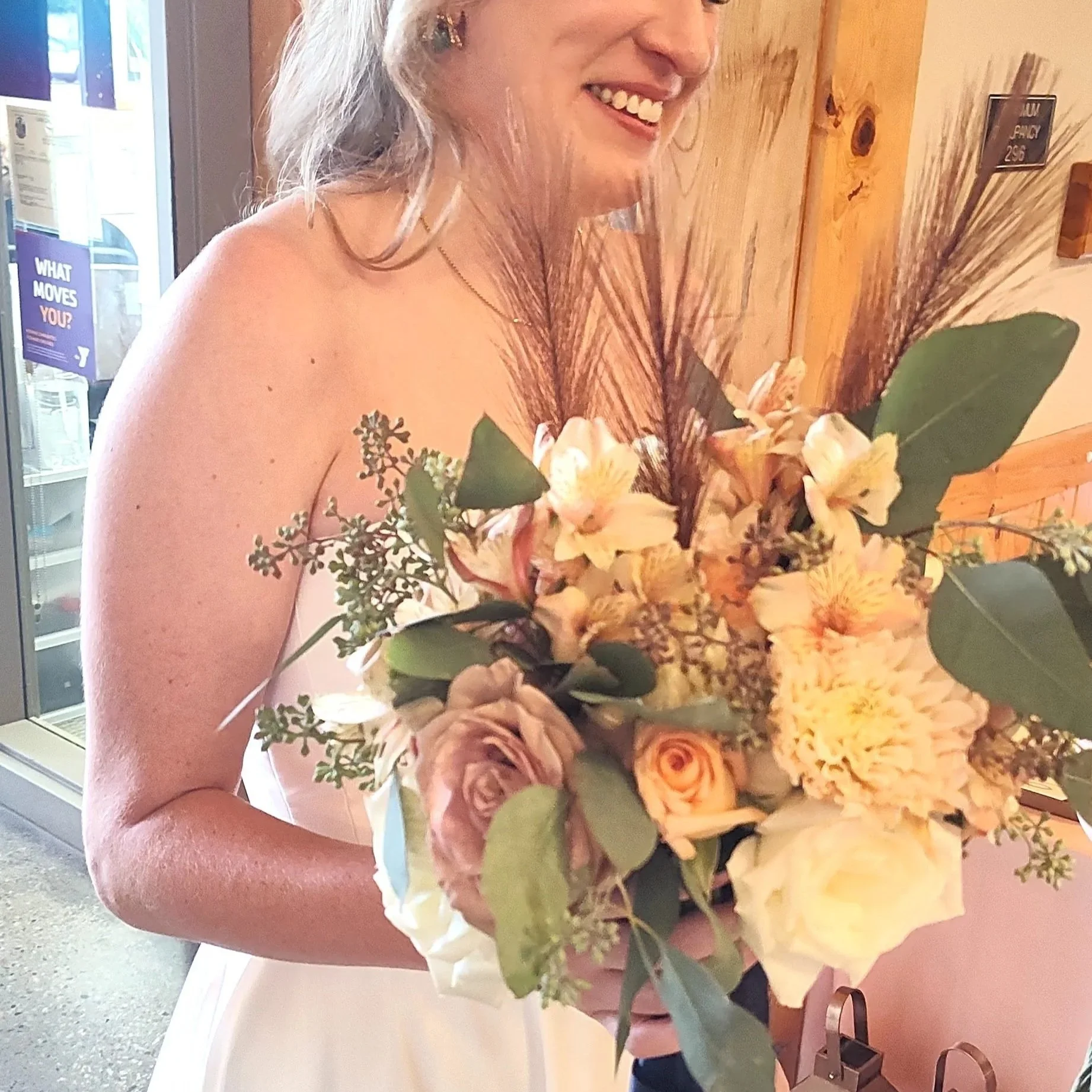 A woman in a sleeveless dress holding a large bouquet of flowers, smiling, with a wooden wall and a door with a purple sign in the background.