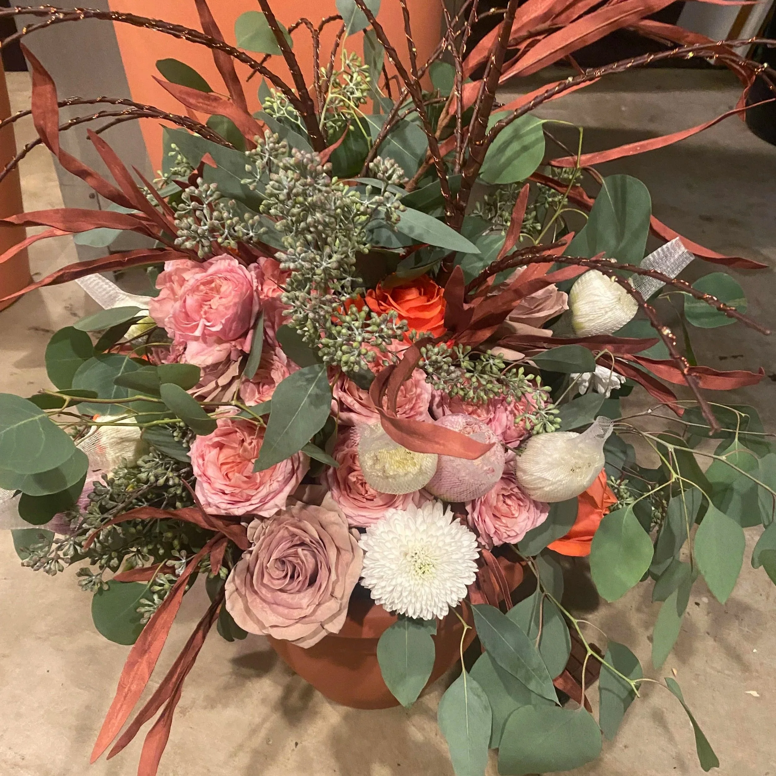 Arrangement of pink roses, white chrysanthemums, dried fruits, and various green and reddish leaves in a brown pot.
