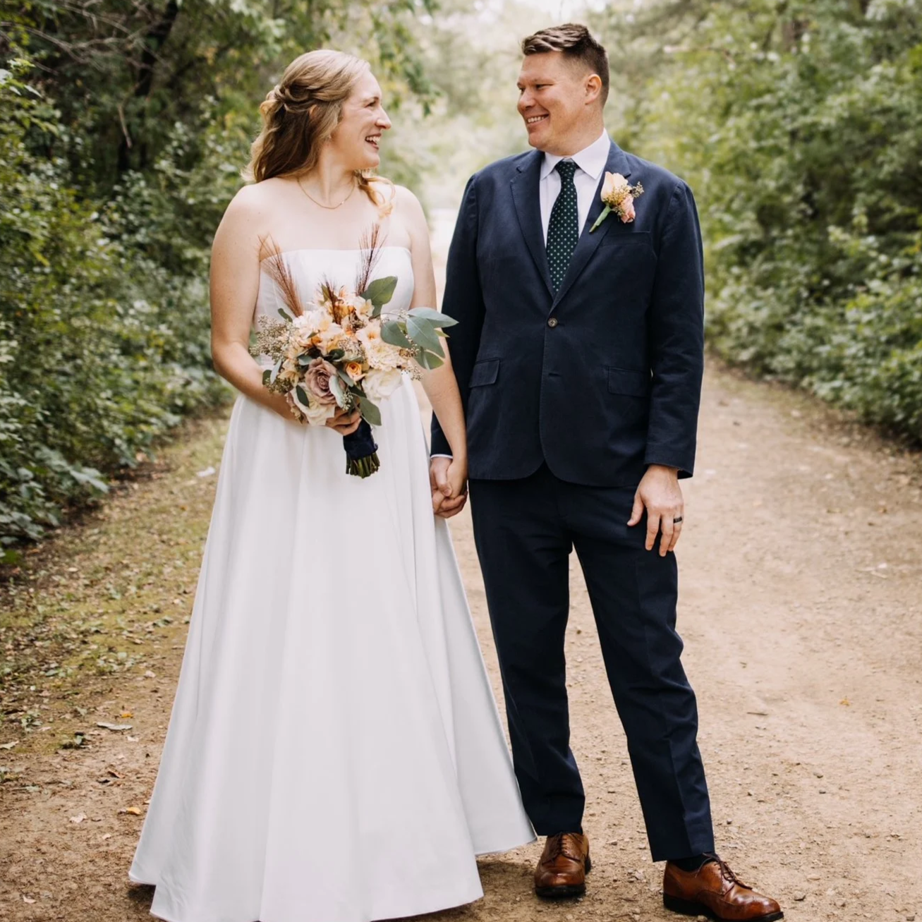 A bride and groom holding hands, standing on a forest path, smiling at each other.