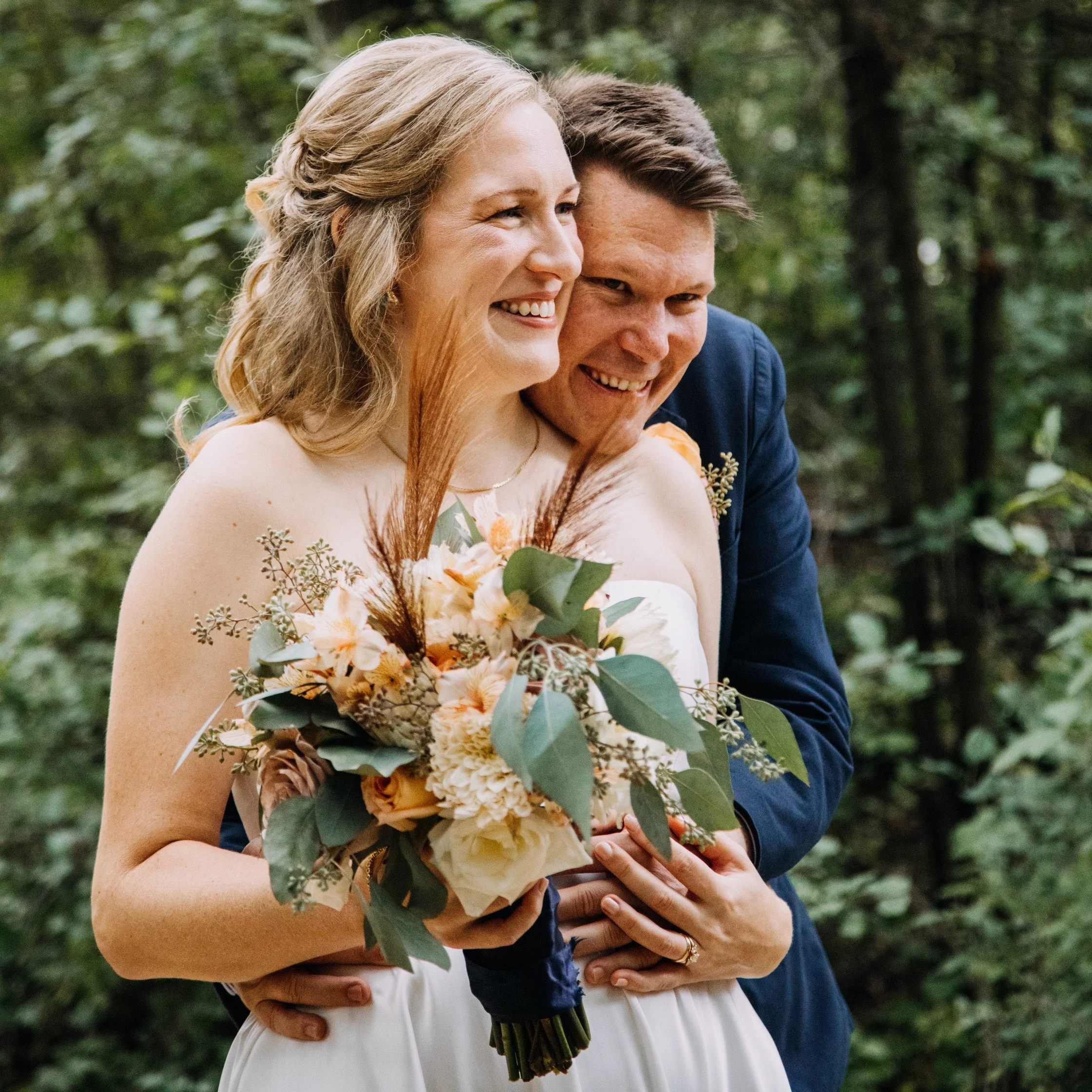 A smiling bride holding a bouquet of flowers in a forest setting, with a groom embracing her from behind.