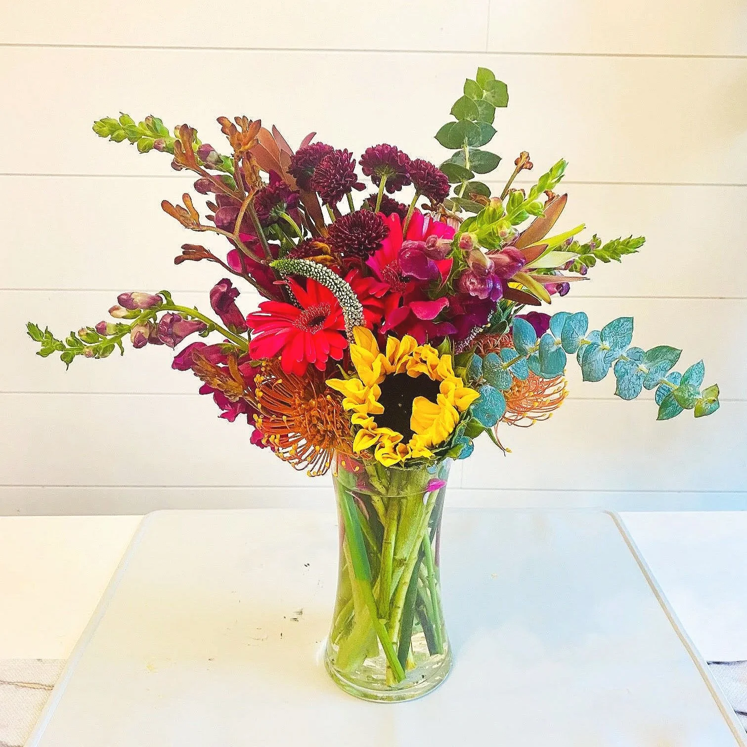 Colorful mixed flower bouquet in a clear glass vase on a white table against a white wall.