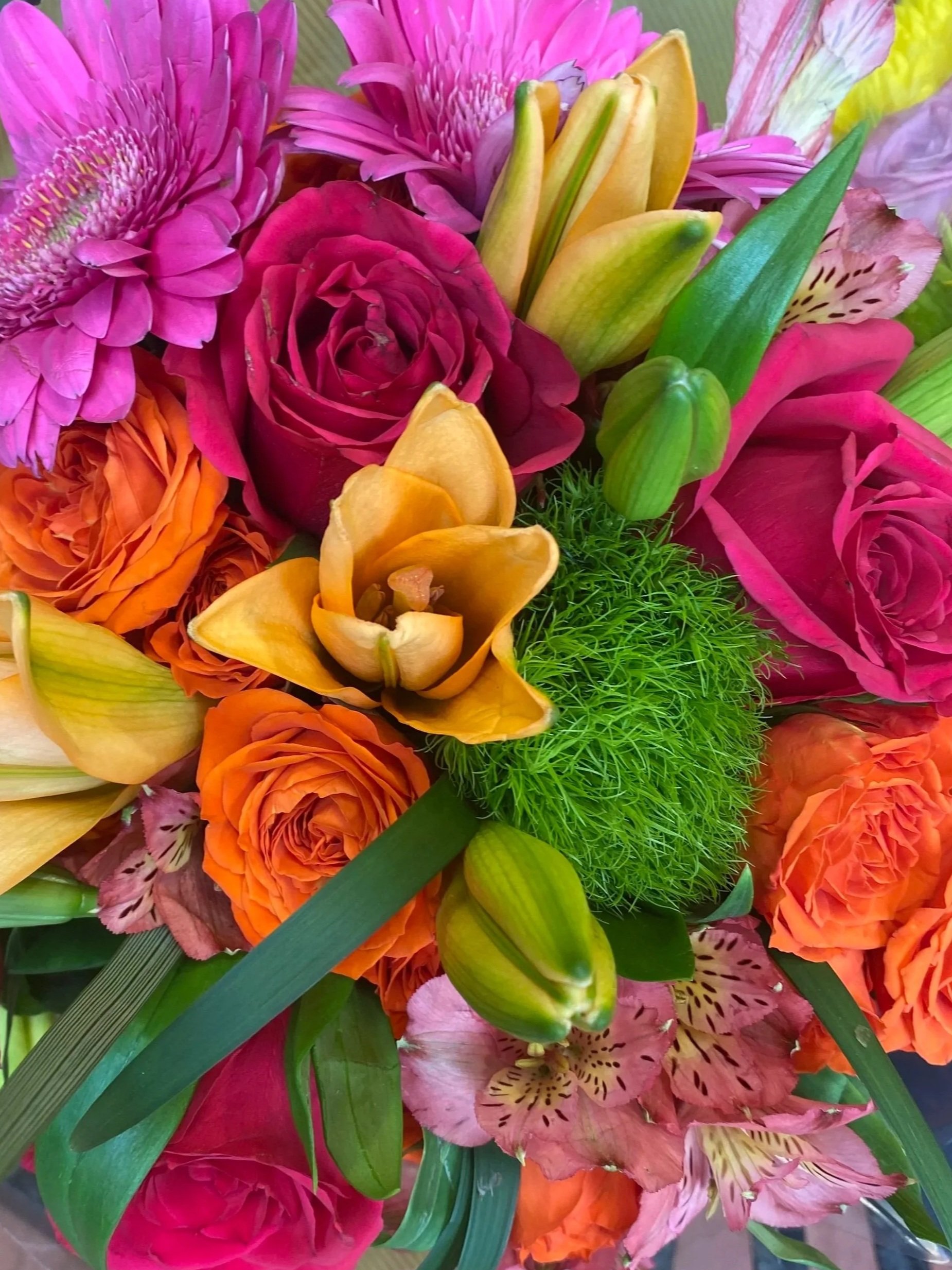 A colorful bouquet of flowers including pink daisies, red roses, orange roses, yellow lilies, and green foliage.